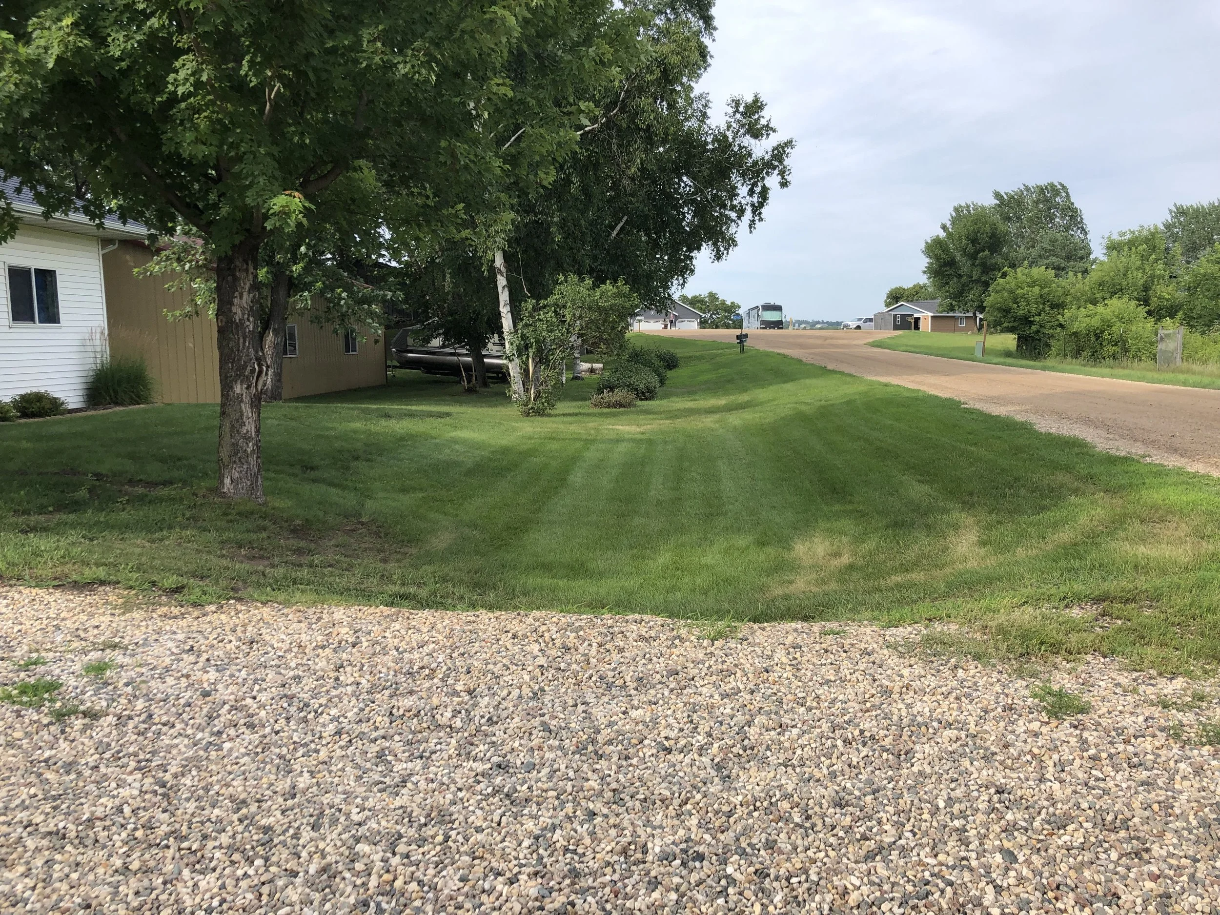 View of a suburban residential area with a gravel driveway, green grass, trees, houses, and a dirt road under a cloudy sky.