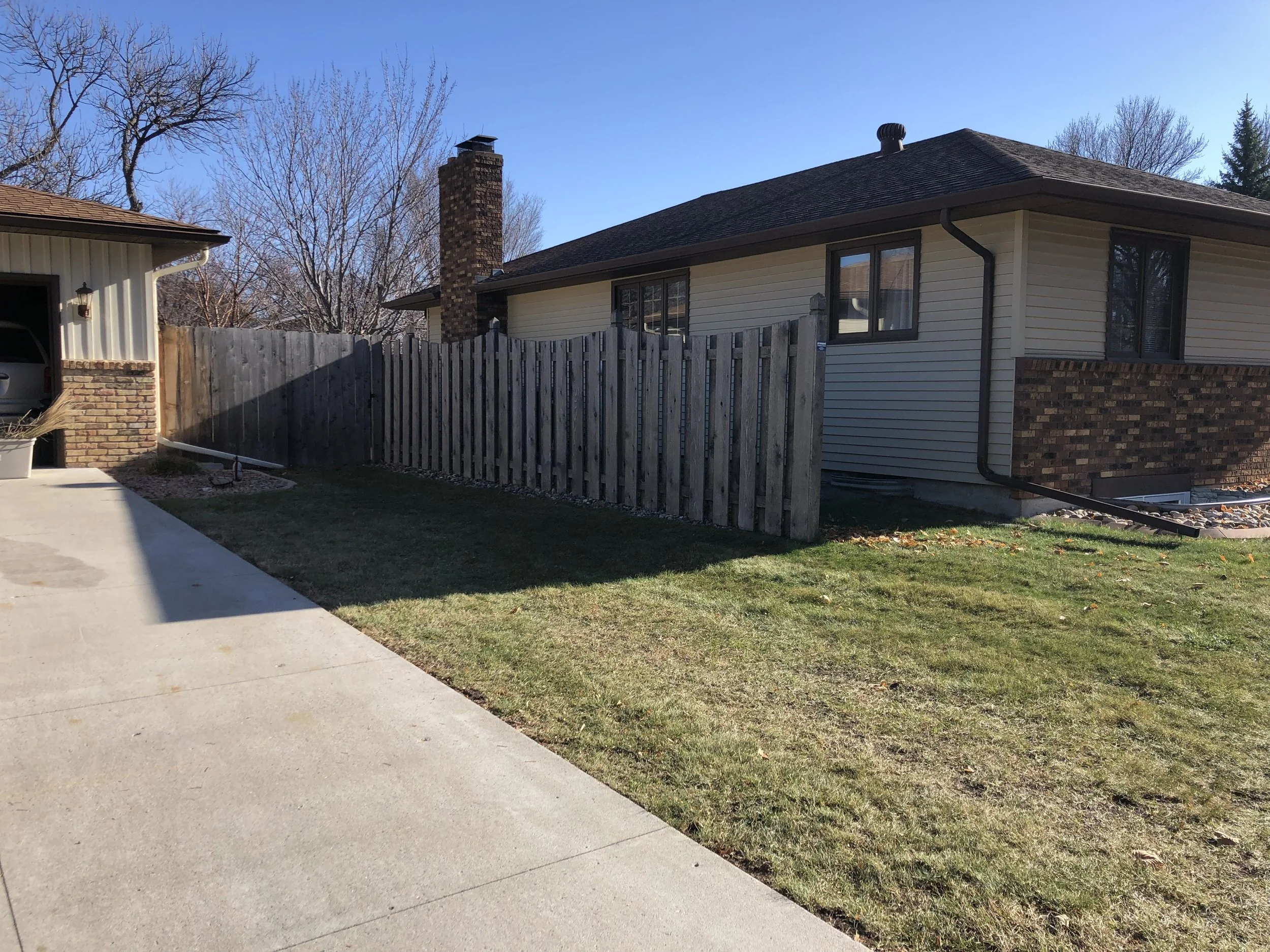 View of a backyard with a wooden fence, a house with beige siding and brick foundation, a chimney, and a concrete driveway under a clear blue sky.