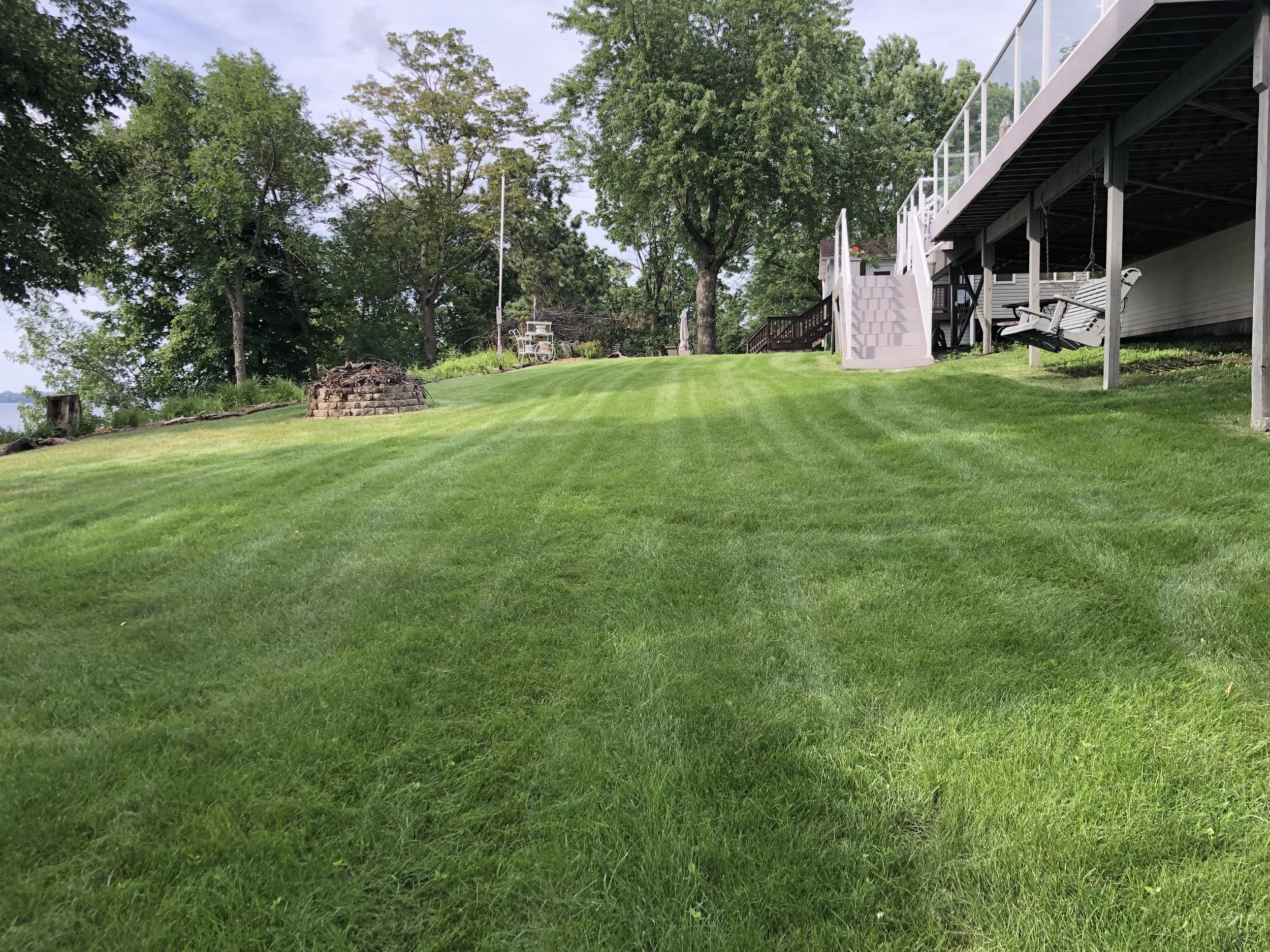 A well-maintained grassy backyard with a staircase leading up to a deck, surrounded by trees and outdoor furniture.