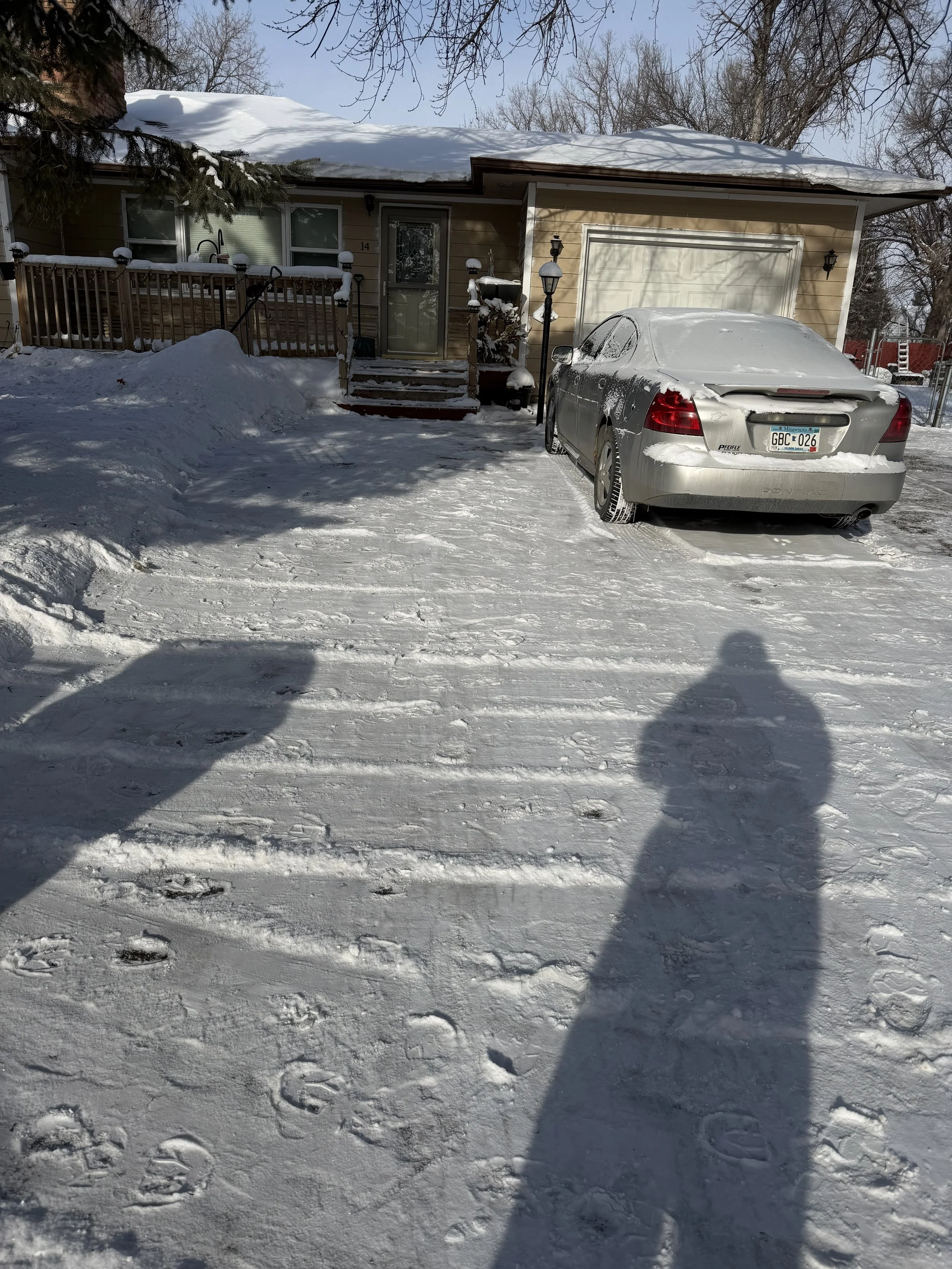 Snow-covered front yard and driveway of a house with a gray car parked in front, partially covered in snow, under a snow-laden roof. A fence and stairs lead to the front door, with outdoor lampposts and snow piles visible.