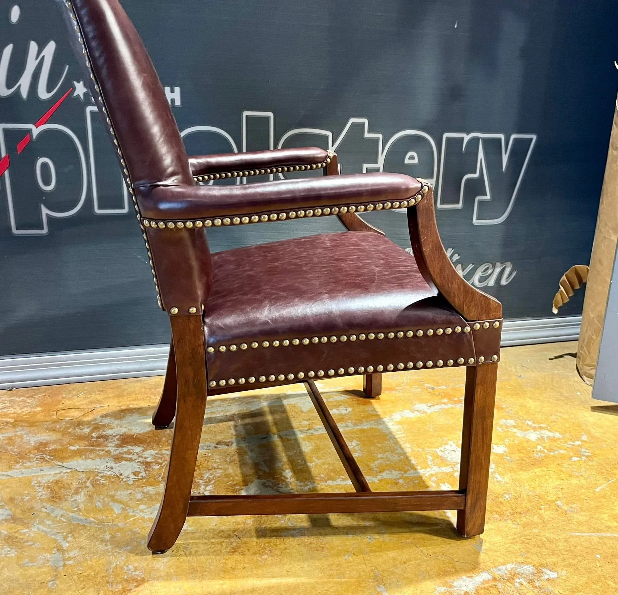 A vintage wooden armchair with leather upholstery and brass nailhead trim, positioned on a worn wooden floor, with a black sign in the background reading 'Furniture'.