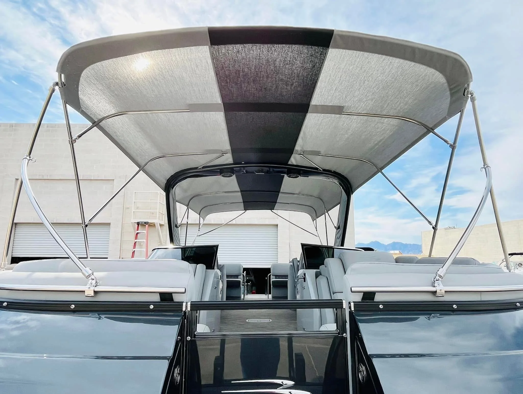 Front view of a modern luxury boat or yacht with a large canopy, white seating, and a clear windshield, docked at a marina with a building and mountains in the background.