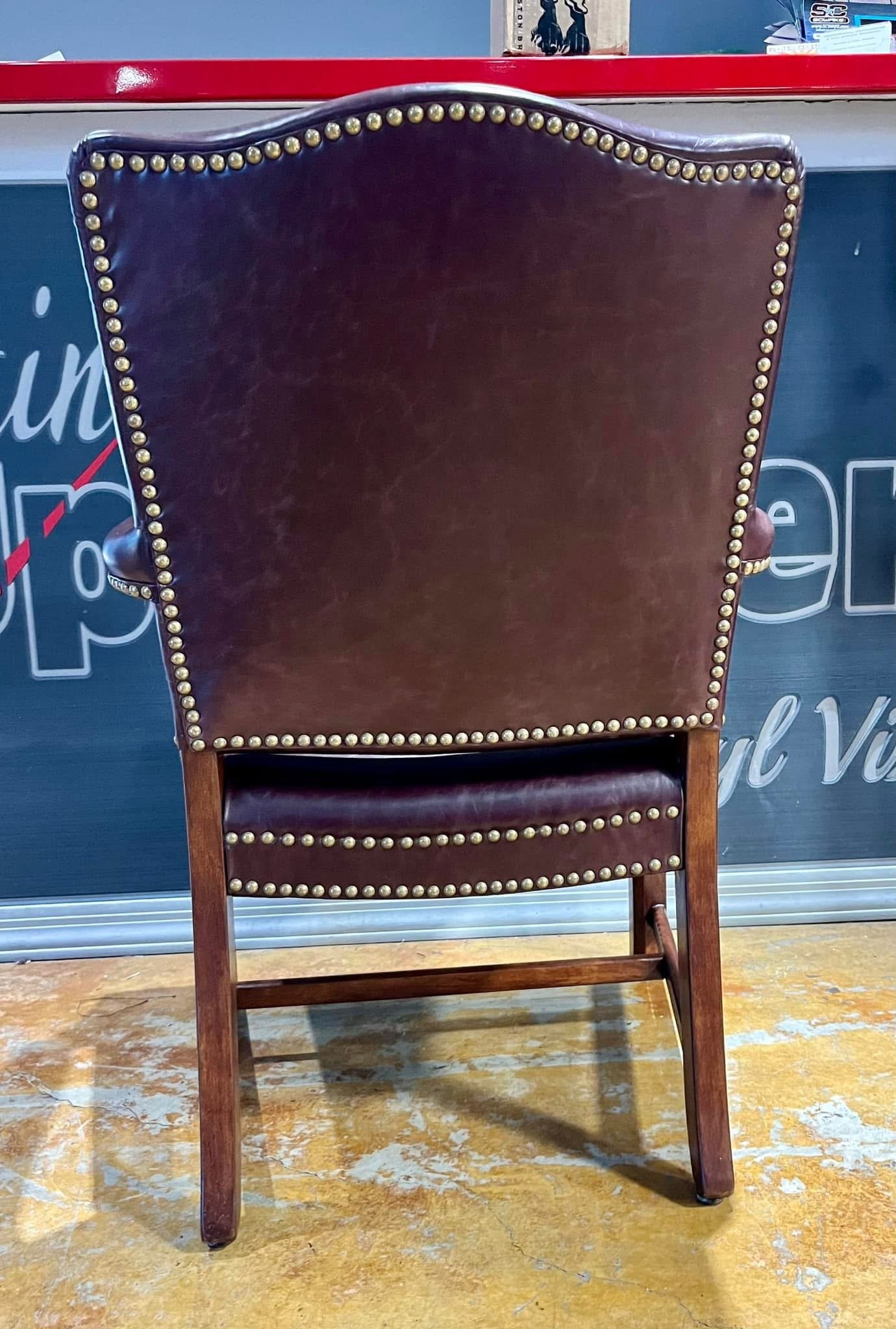 Back of a vintage leather armchair with brass nailhead trim, standing on a worn wooden floor.