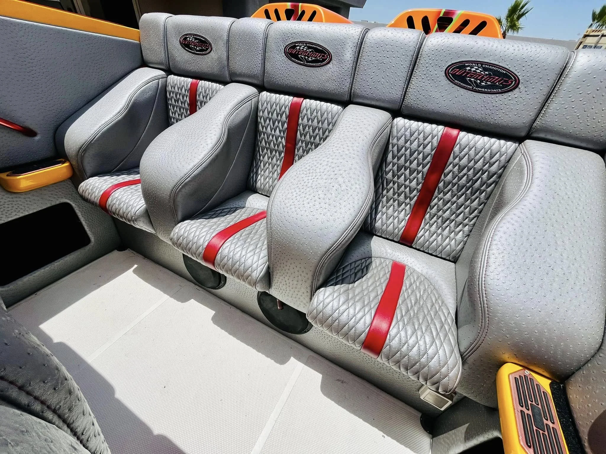 Gray boat seating with red seatbelts and custom cushions, in an outdoor setting with orange accents and palm trees in the background.