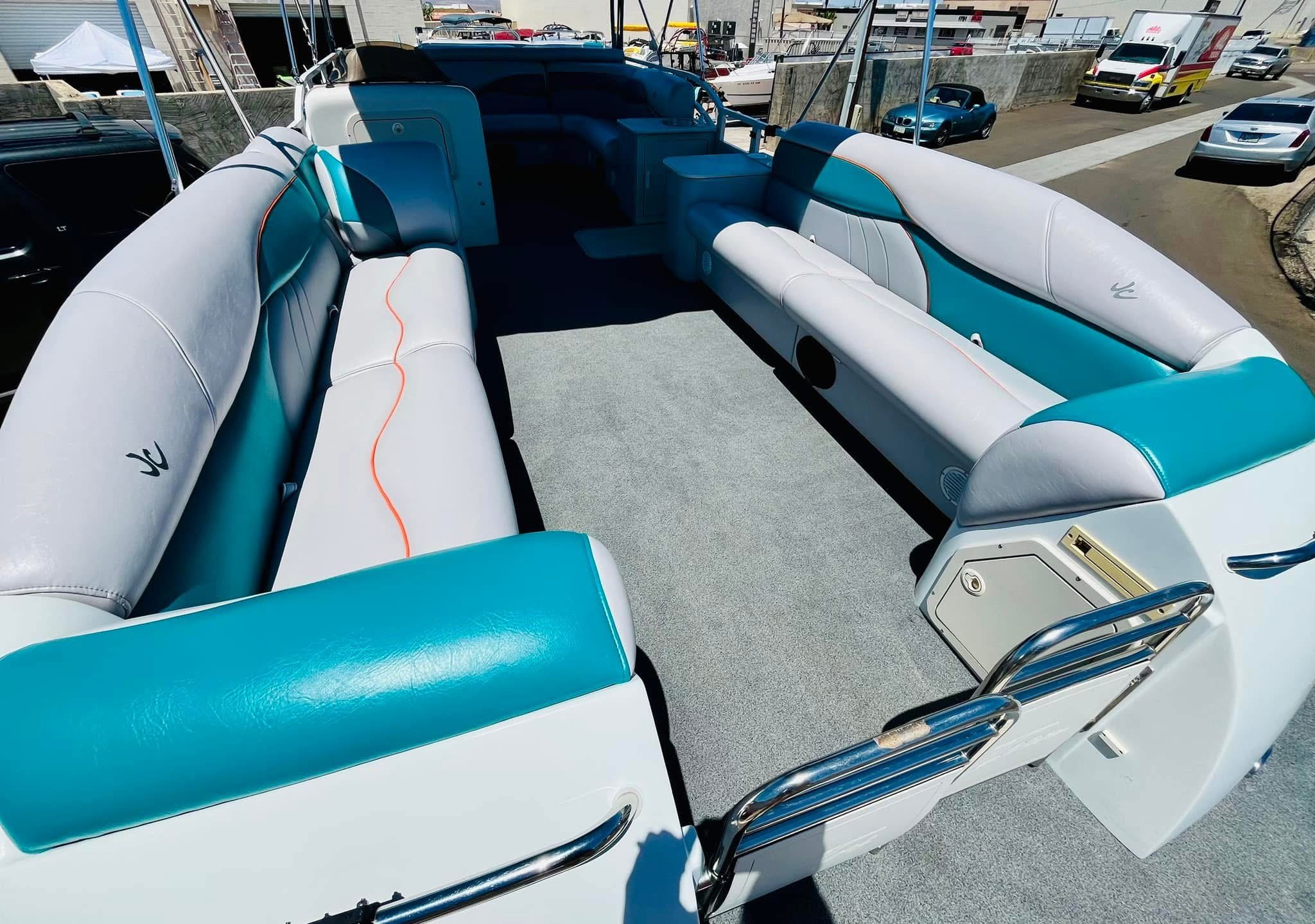 Interior of a white and turquoise pontoon boat with padded seating, a carpeted floor, and outdoor parking lot with several cars and vans in the background.