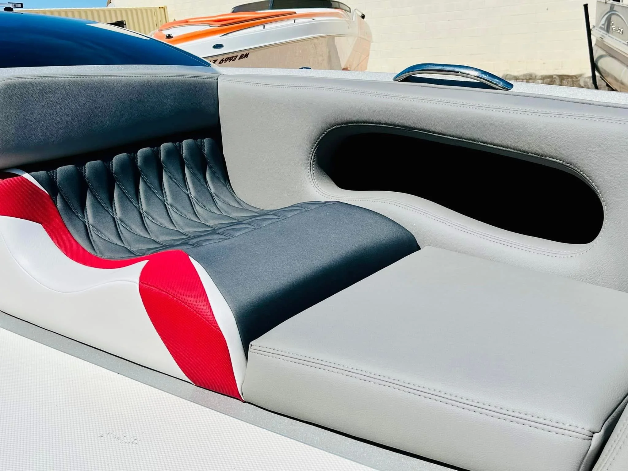 Close-up of the seat and interior side of a boat with white, red, and dark gray upholstery, with boats and water in the background.