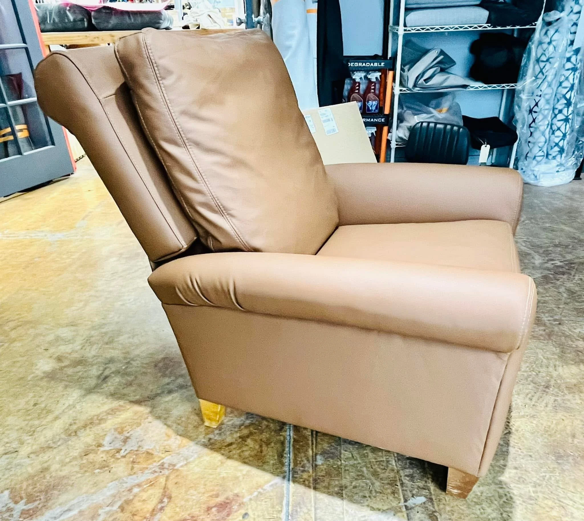 A tan leather high-back armchair with a cushioned headrest, positioned on a multicolored tile floor in a room with shelves and clothes in the background.