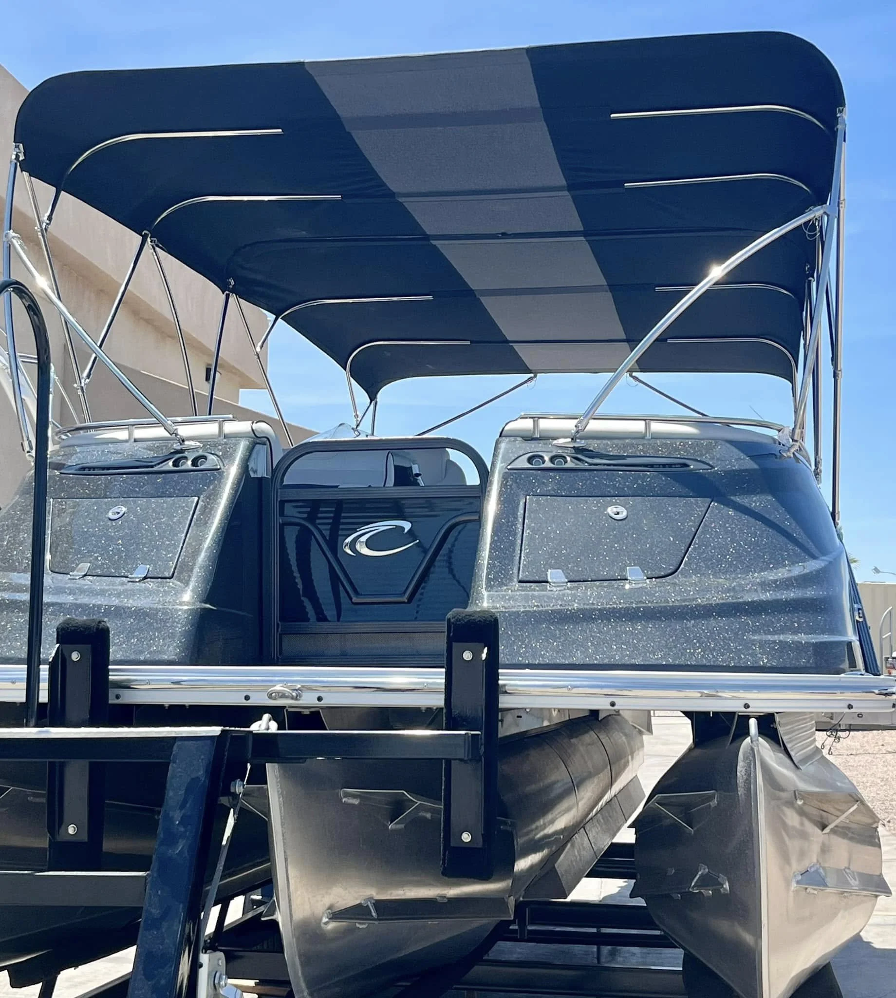 A pontoon boat with black and gray speckled exterior, a canopy, and metal pontoons, parked on a trailer outdoors under a blue sky.