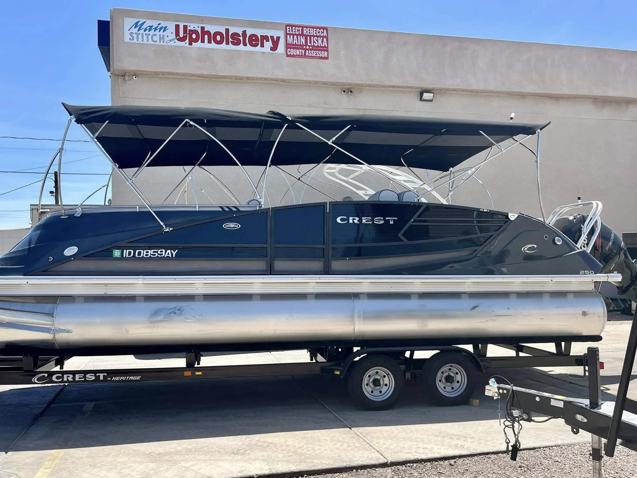 A Crest pontoon boat parked on a trailer in front of a beige building with a sign for upholstery services and a political campaign sign. The boat has a black and silver color scheme with a canopy, and the trailer is labeled Crest Heritage.