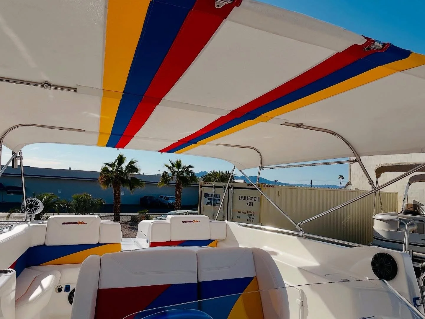 View of a boat's interior with colorful striped fabric on the ceiling, white seating with red, blue, and yellow accents, and palm trees and mountains in the background.