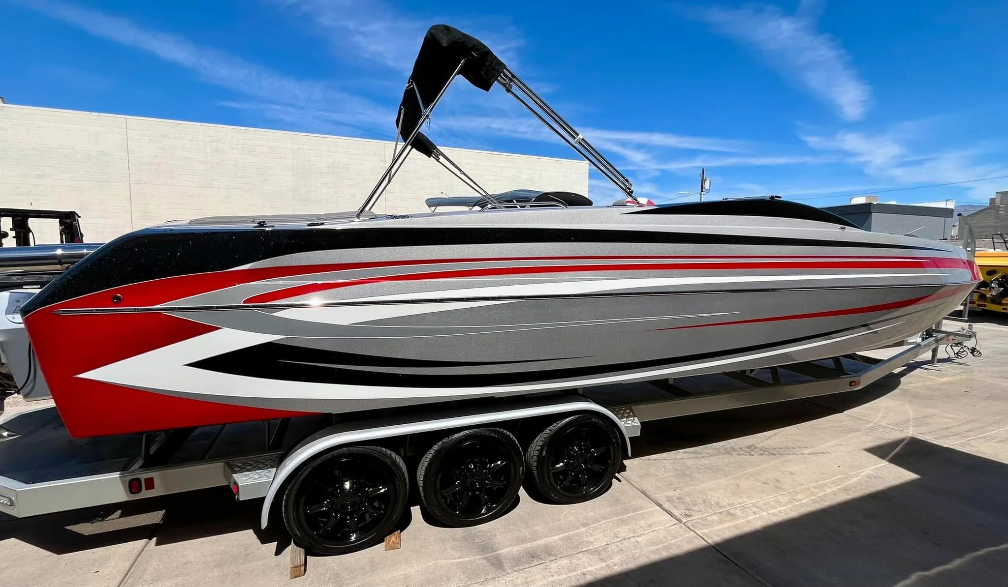 A sleek speedboat with a black, red, white, and gray design, mounted on a trailer with three black wheels, parked outdoors under a blue sky.
