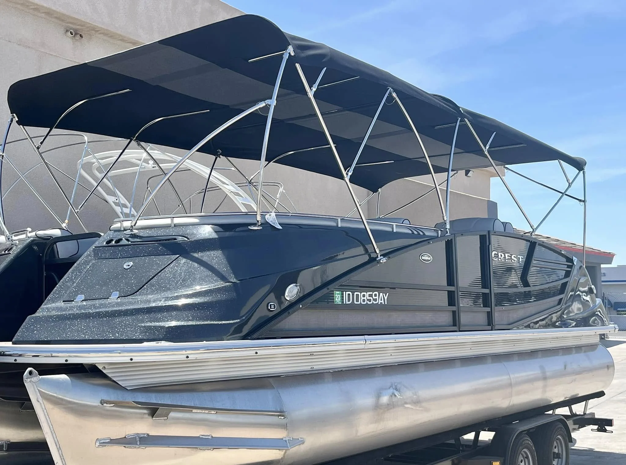 A black pontoon boat on a trailer with a blue canopy, parked outdoors near a building.