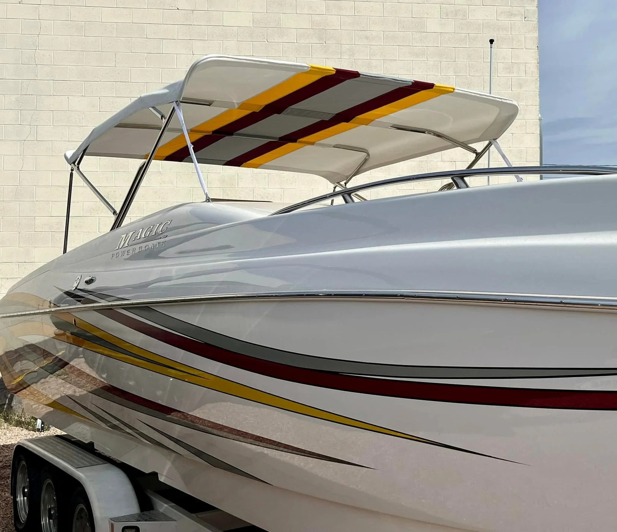A white powerboat with red, black, and yellow stripe decals on a trailer against a brick wall with partially cloudy sky.