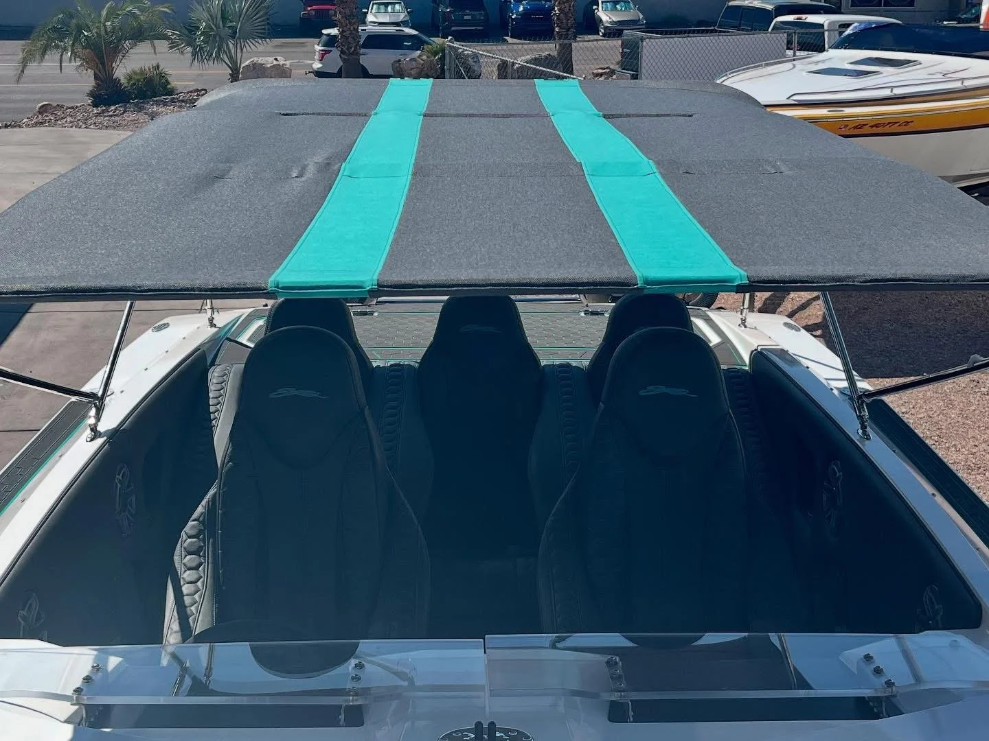 A boat with a black canopy and blue straps over the top, showing four black seats inside, parked at a marina with other boats and parked cars in the background.
