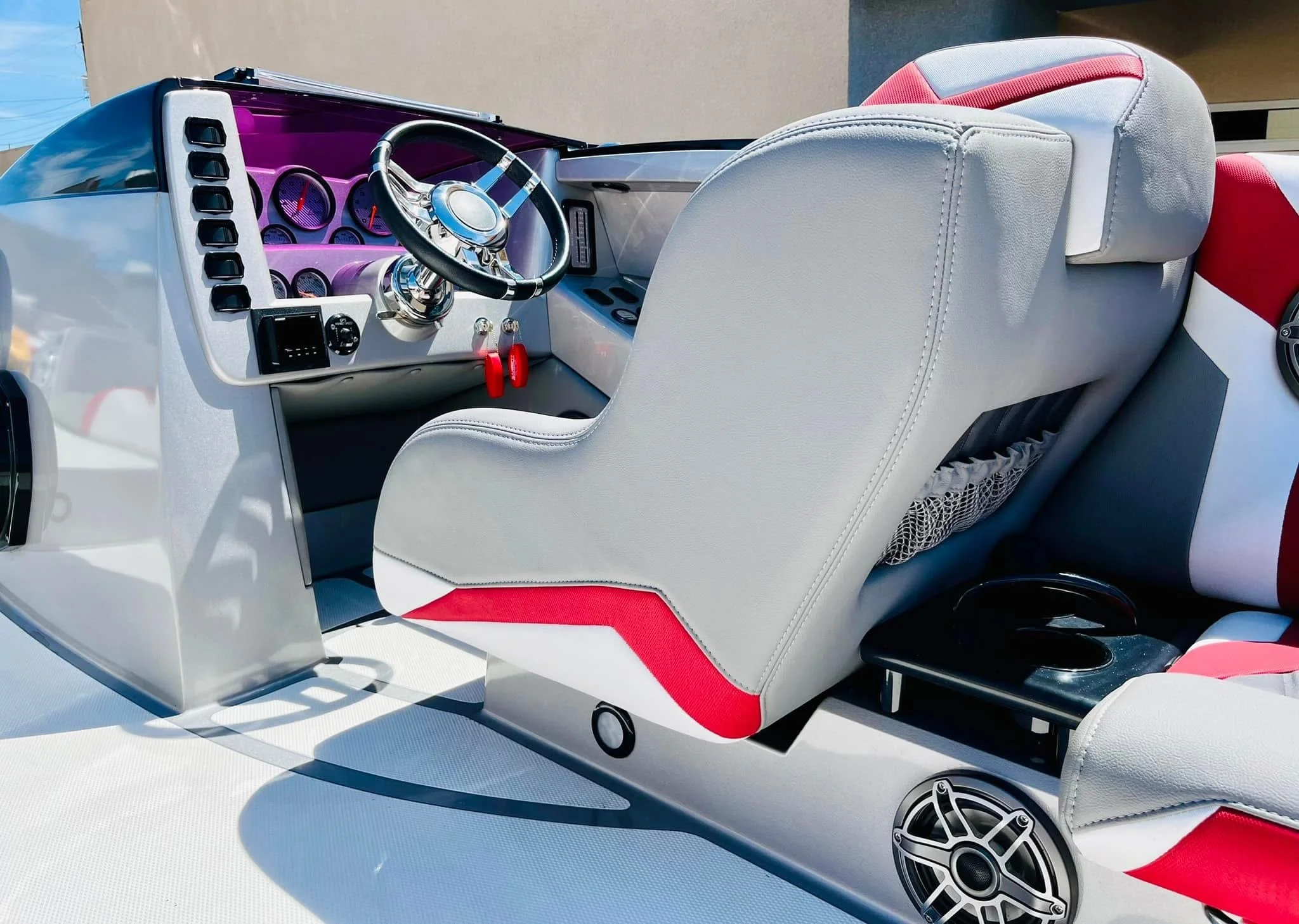 Close-up of a boat's helm showing a white and red captain's seat, dashboard with multiple gauges, steering wheel, and speakers.