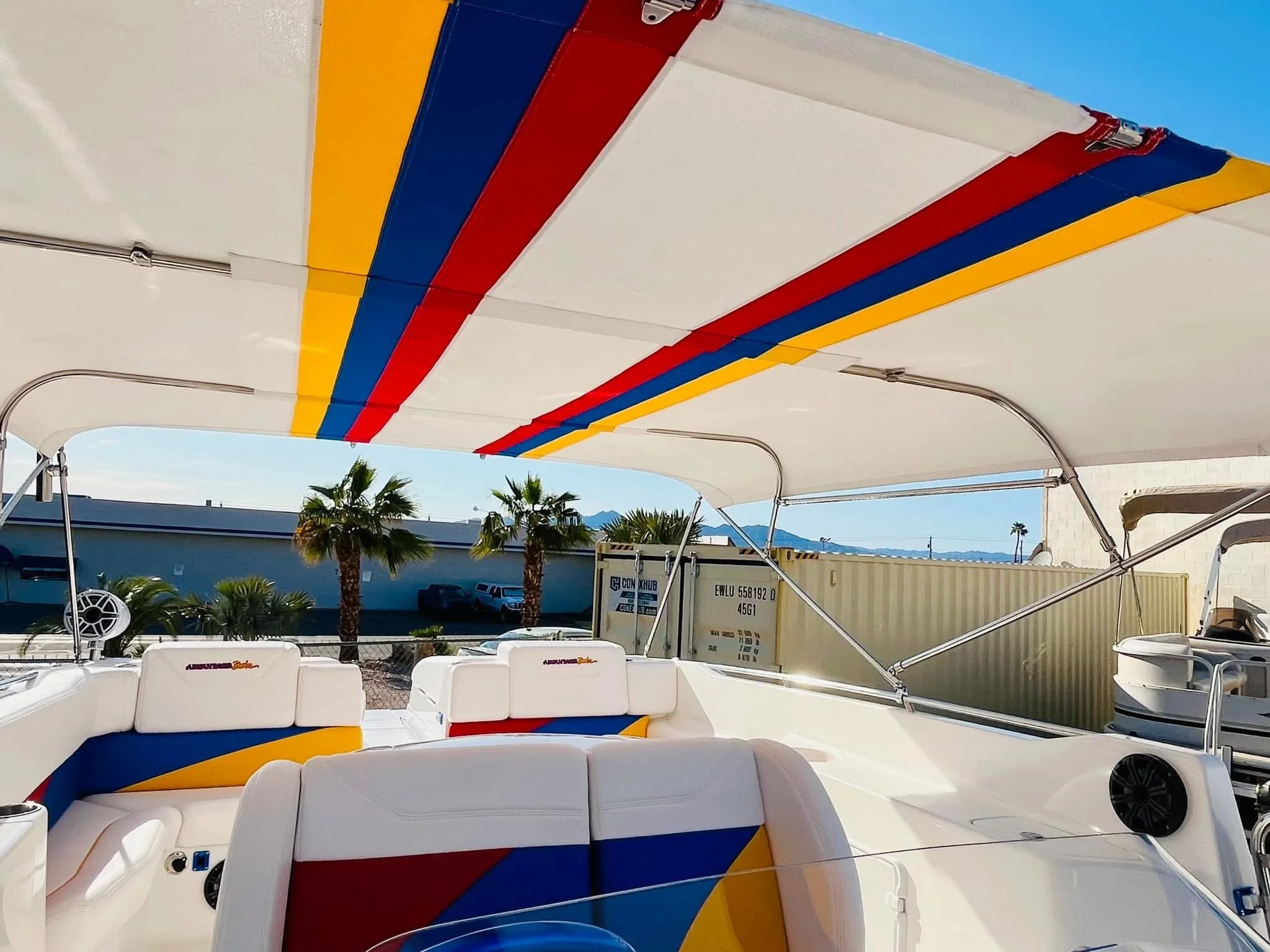 The image shows the interior of a boat with a white canopy featuring colorful red, blue, and yellow stripes. There are white cushioned seats with matching colorful patterns, a small black speaker, and a sunshade with palm trees and a clear blue sky v