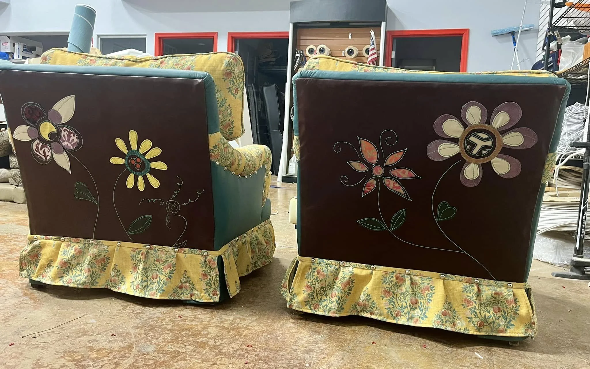 Two upholstered chairs with floral patterns and decorative floral designs painted on the back, in an indoor space with shelves and equipment in the background.