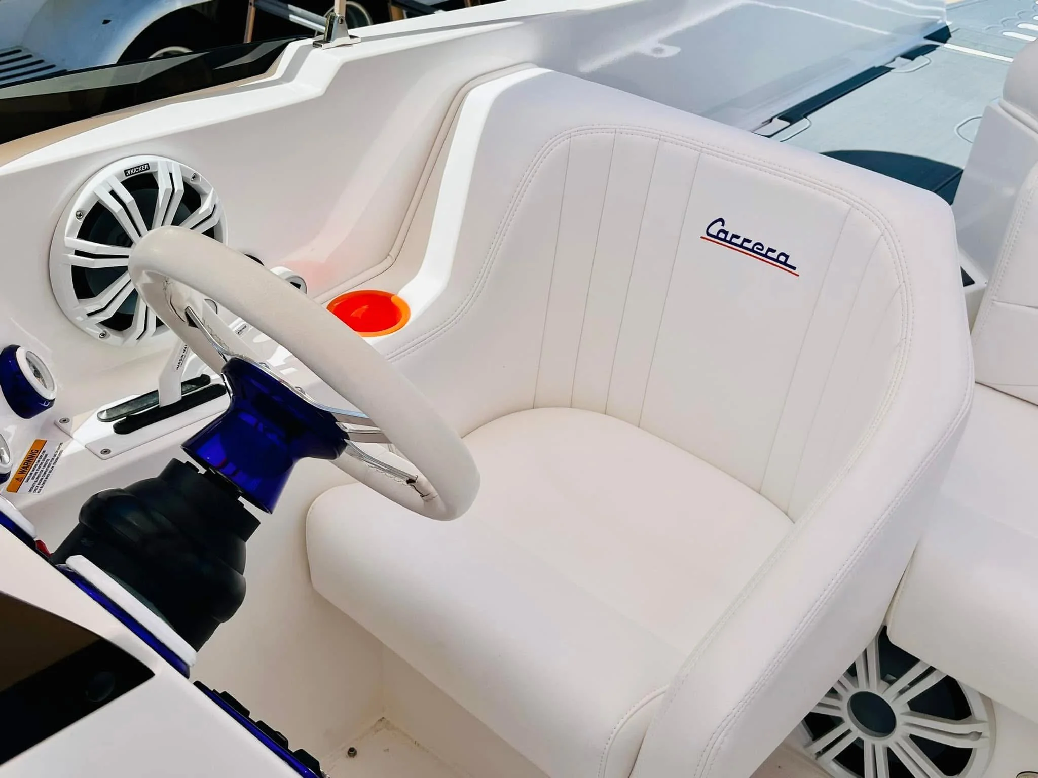 Close-up of a white boat interior featuring a white cushioned seat with the Carrera logo embroidered on the backrest, a white steering wheel with a blue hub, circular white speakers, and an orange cup holder.