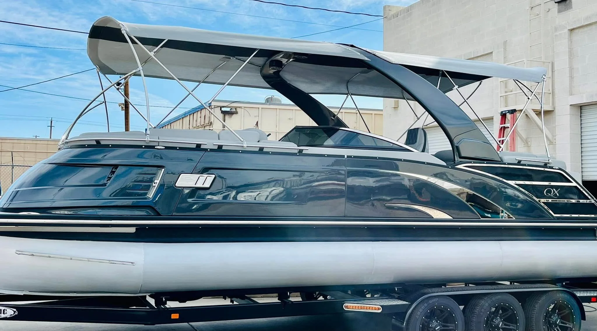 A luxury motorboat is on a black trailer with six wheels, parked outdoors in front of buildings and a blue sky.