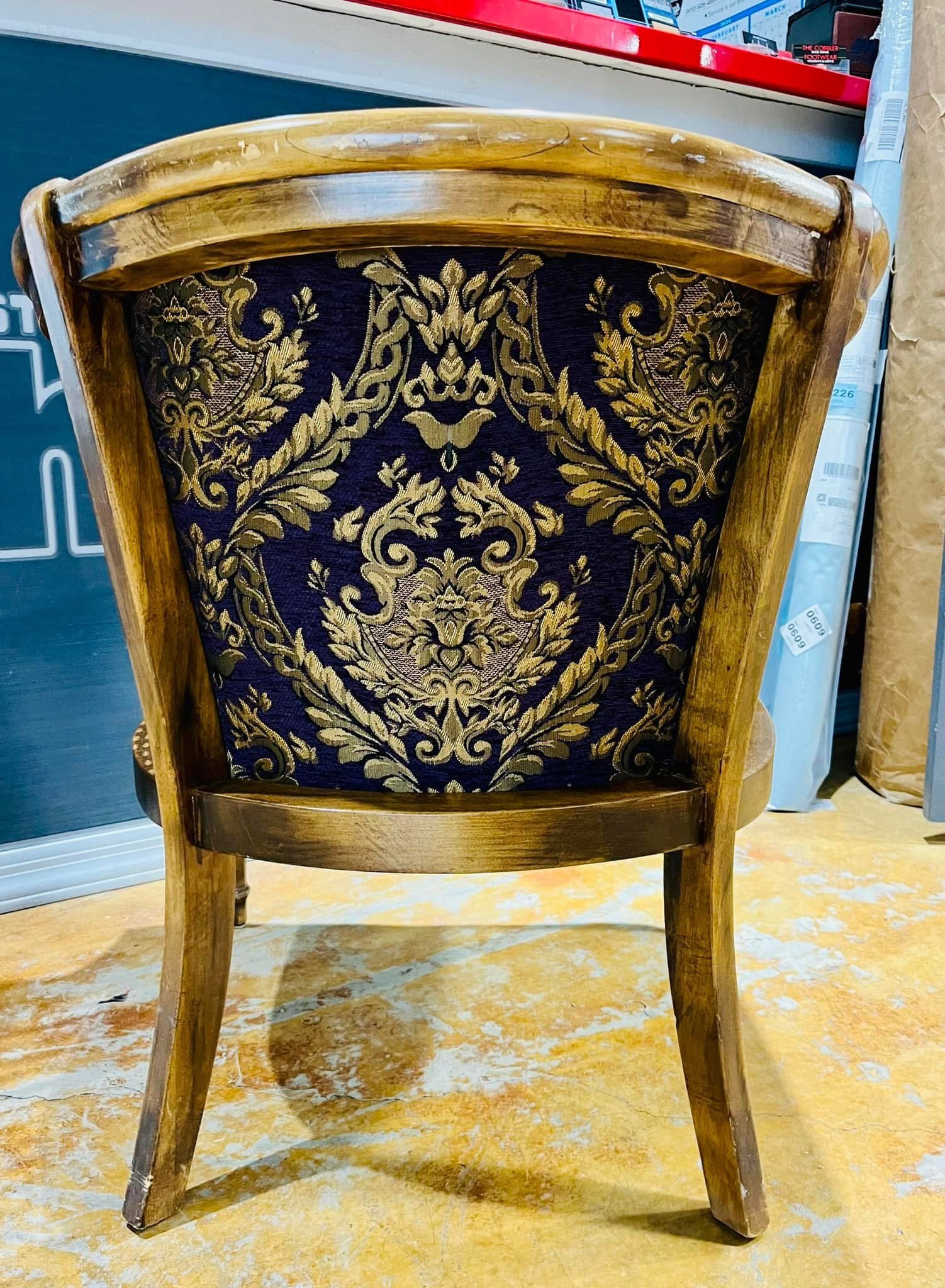 A wooden chair with an intricately patterned navy and gold upholstered backrest, placed on a worn yellowish floor.