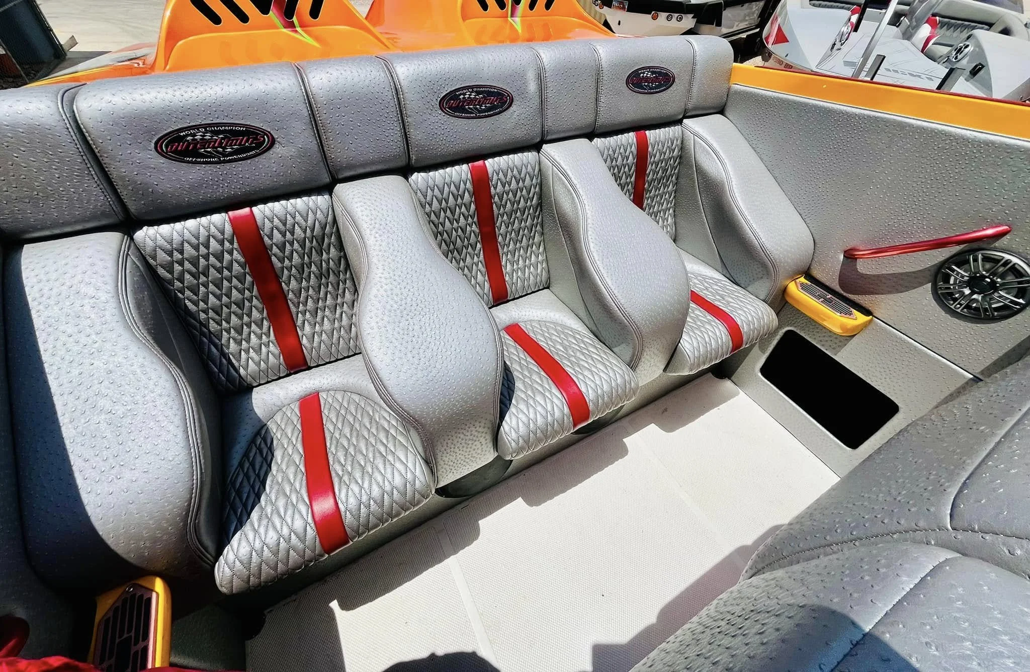 Close-up of a boat's interior seating area, featuring three silver diamond-stitched seats with red accents and speaker, with boat exterior visible at the back.