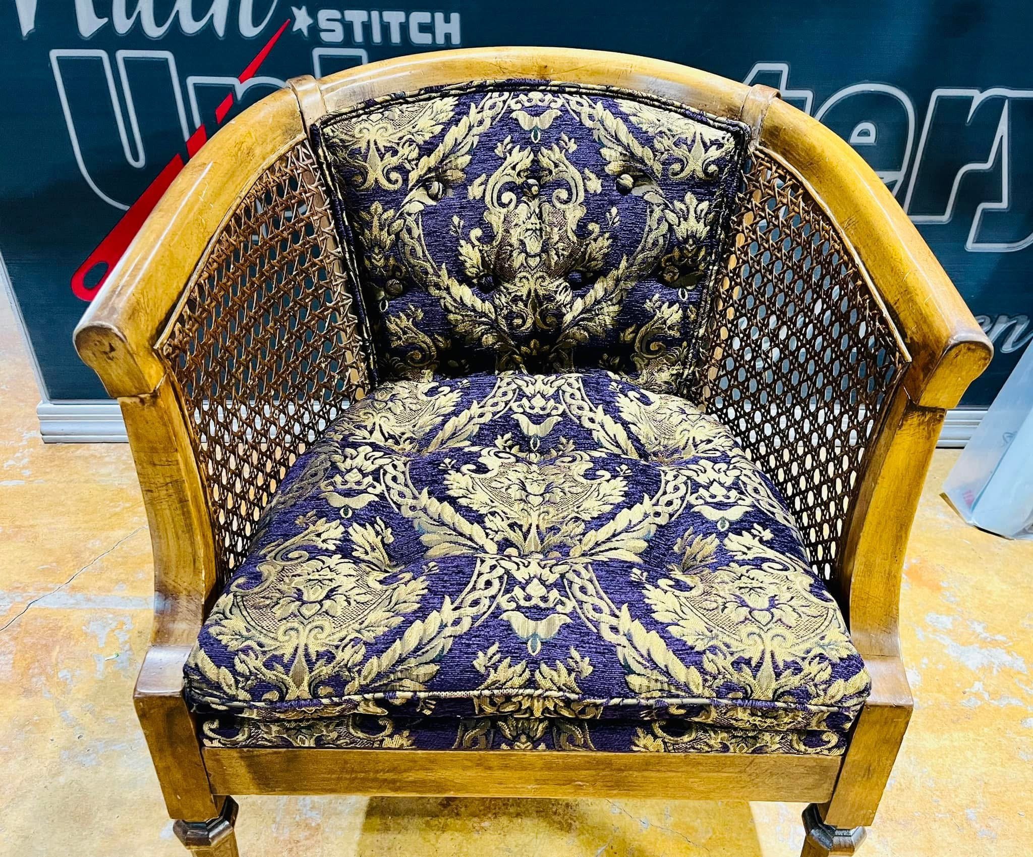 A vintage wooden armchair with a rounded back and caned sides. The seat and back are upholstered with dark fabric featuring an ornate gold floral pattern.