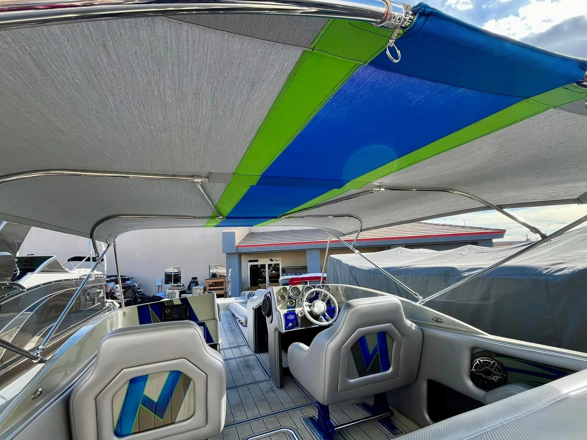 View of boat's cockpit with steering wheel, dashboard, and two white seats, covered by a fabric canopy with blue and green accents, with other boats and a building in the background under partly cloudy sky.