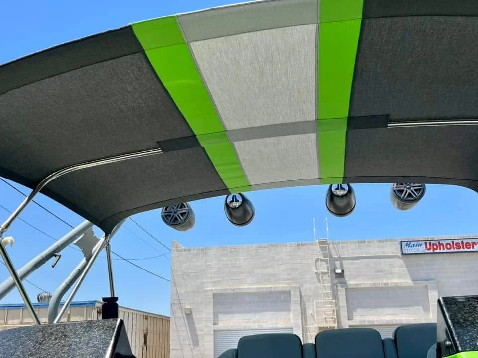 Boat canopy with gray and green fabric, four speakers hanging underneath, and a building with a sign that reads "Main Upholstery" in the background.