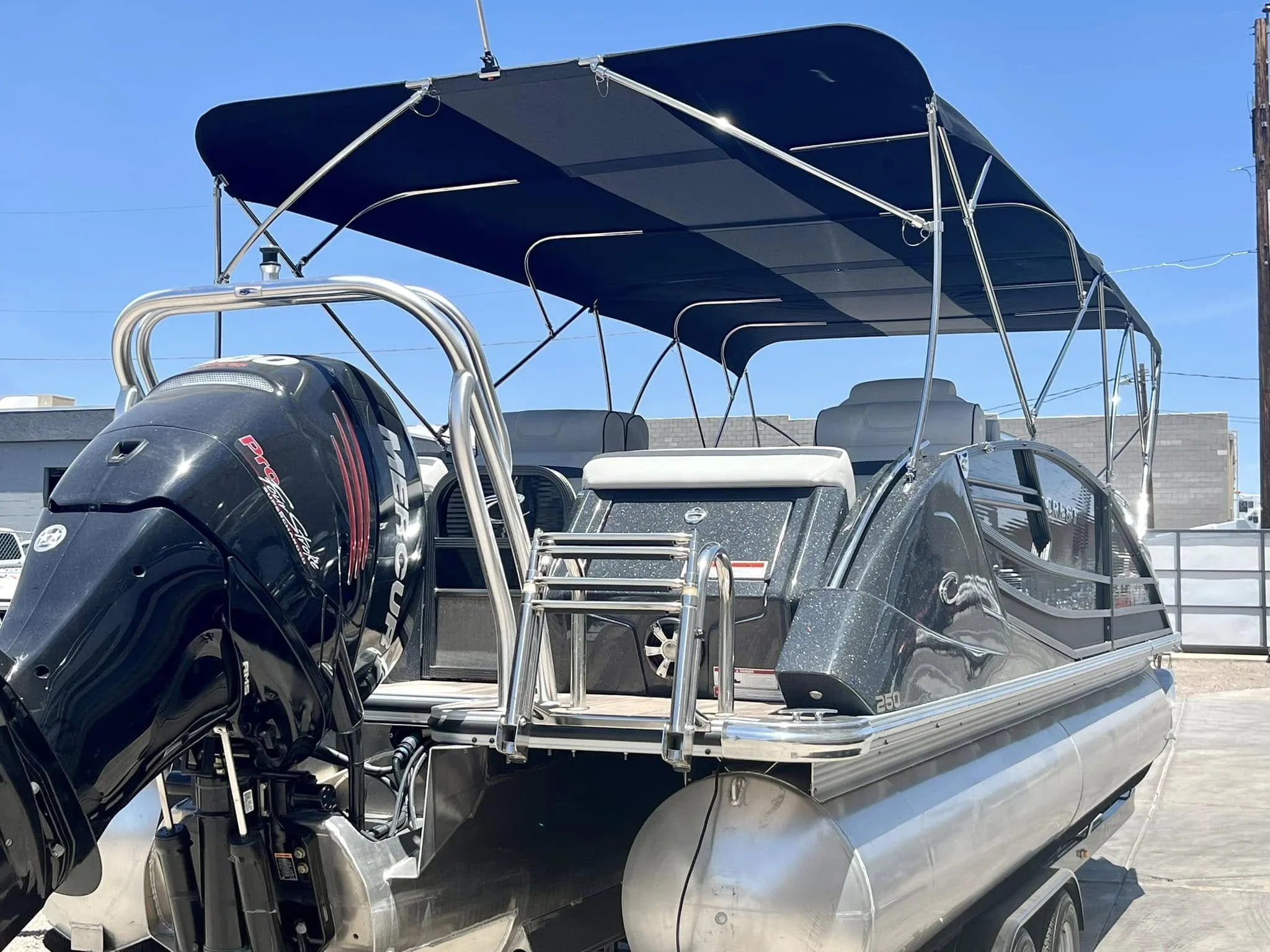 A pontoon boat with a black canopy and a Mercury outboard motor, parked on a trailer against a backdrop of clear blue sky and some buildings.