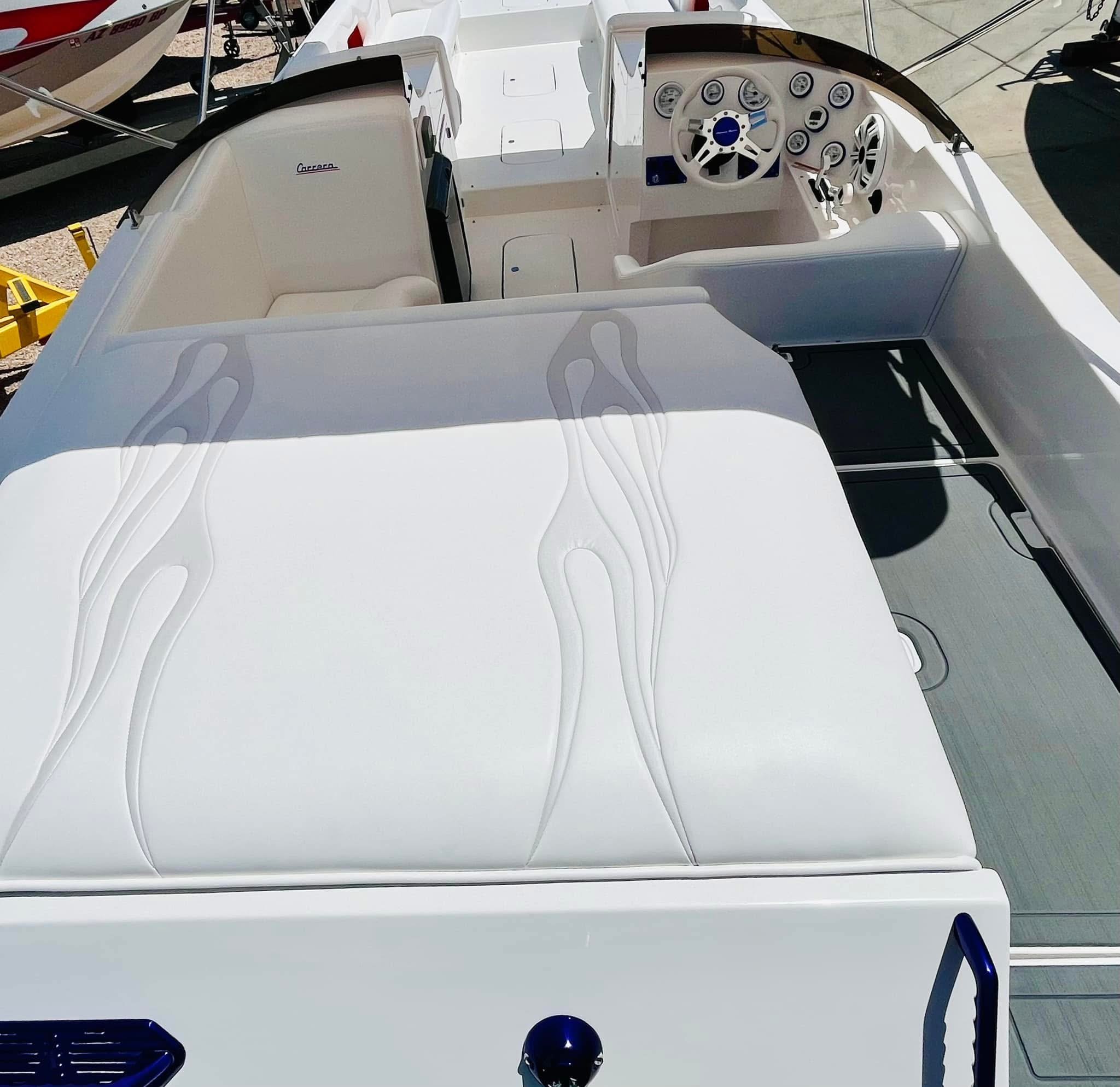 Top view of a white motorboat's deck showing a cushioned sunbathing area, the boat's steering wheel and control panel in the cockpit, and surrounding dock area with other boats.
