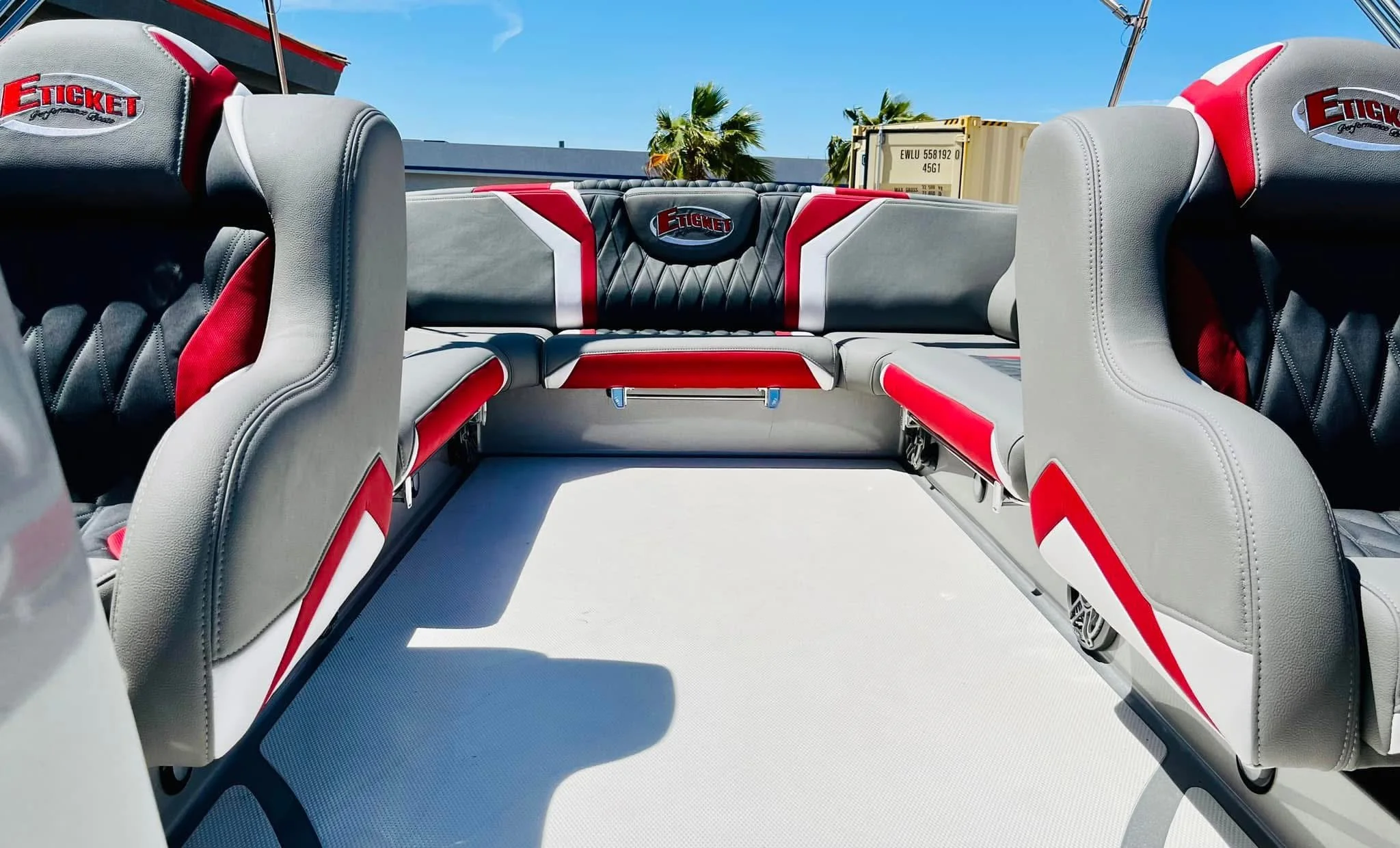 View of boat seats with gray, black, white, and red upholstery, branded with 'Ficket', under sunny skies with palm trees with custom upholstery.