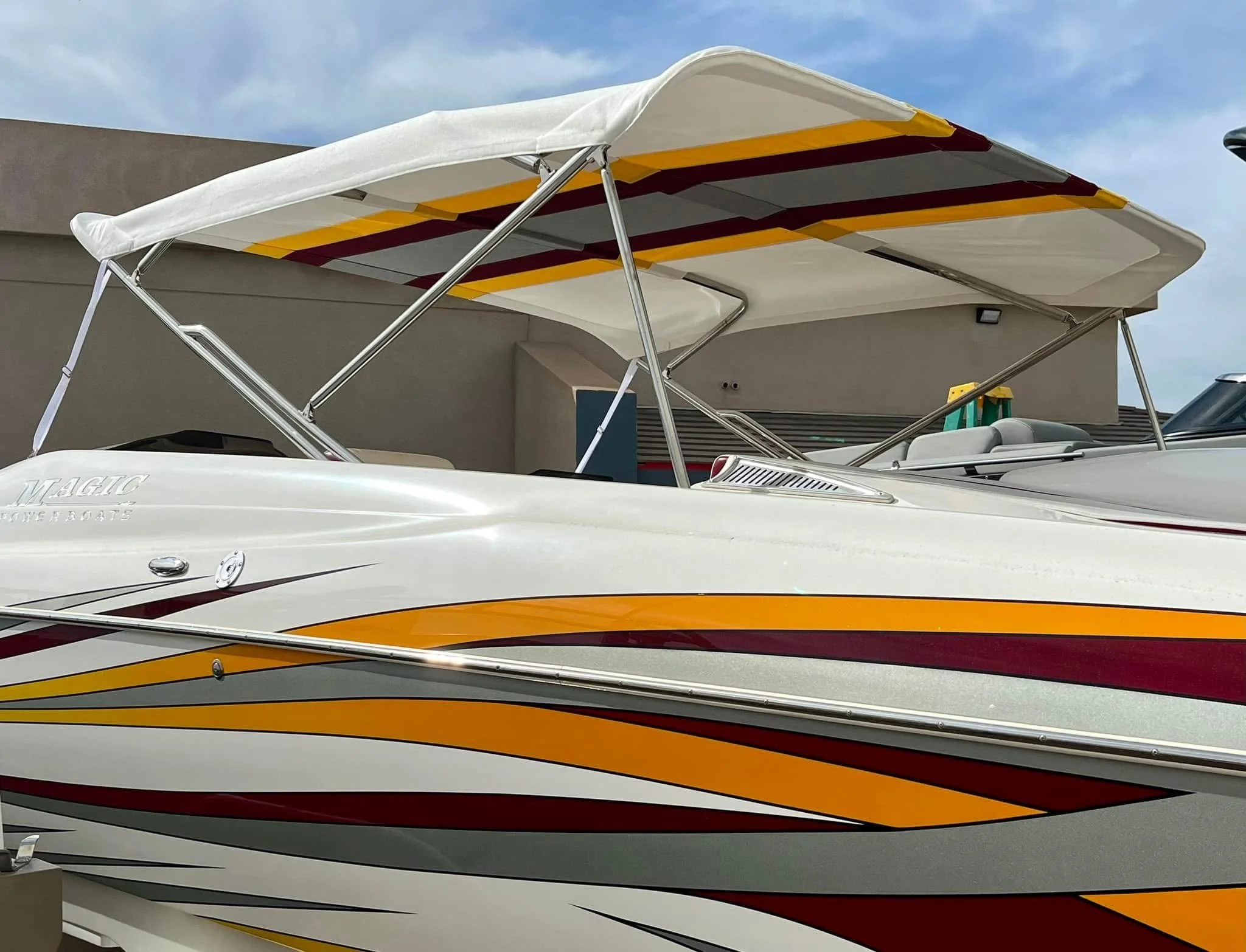 A white powerboat with red, gray, orange, and yellow stripe decals, parked next to a beige building under a partly cloudy sky.