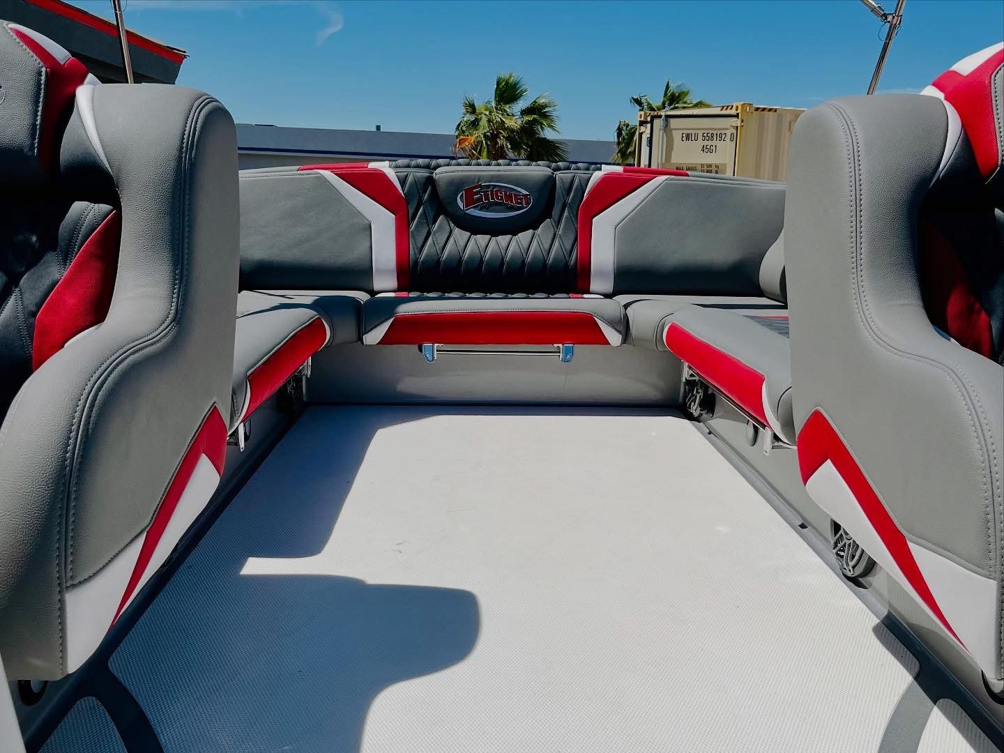 View of the interior of a boat with gray and red cushioned seats and a black, red, and gray backrest with the word 'FIGHTER' on it, under a blue sky with a few palm trees and a shipping container in the background.