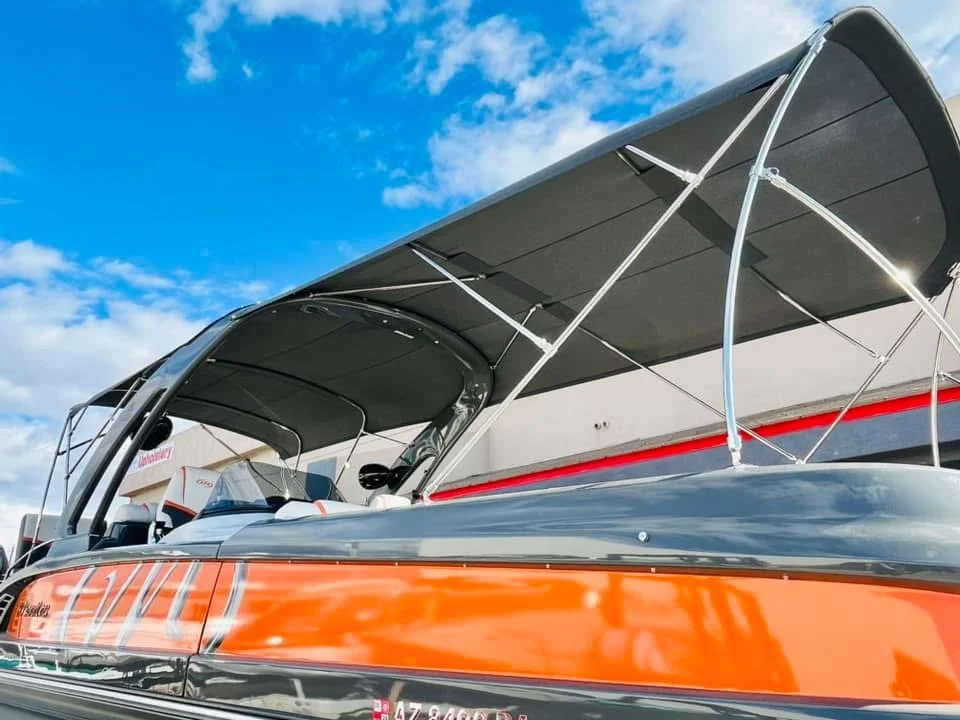 Close-up of a boat docked at a marina, showing the boat's side with an orange and black hull and a black canopy over the seating area, under a blue sky with scattered clouds.