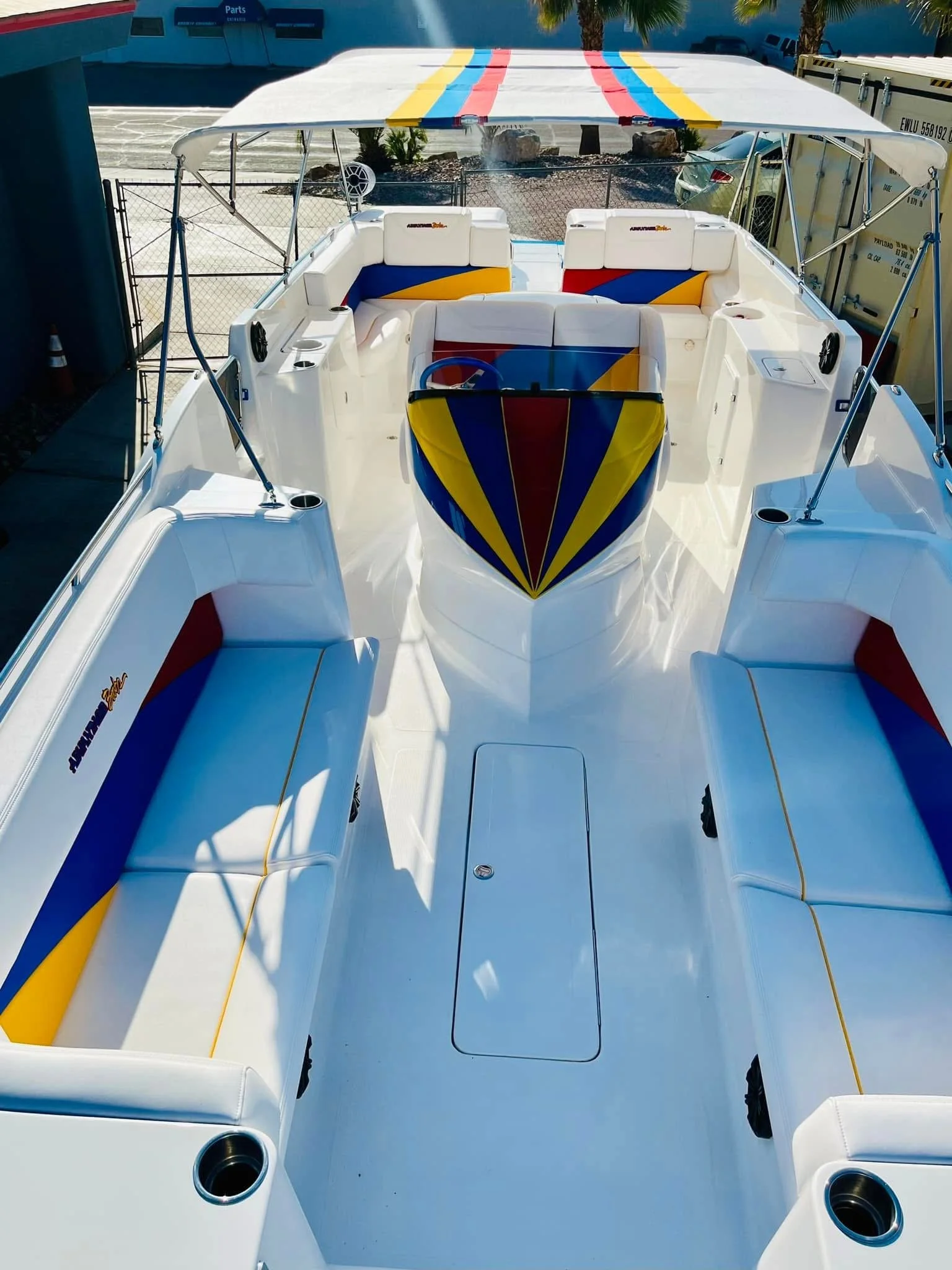 Colorful motorboat with white seating and a rainbow-striped canopy, docked at a marina with palm trees.