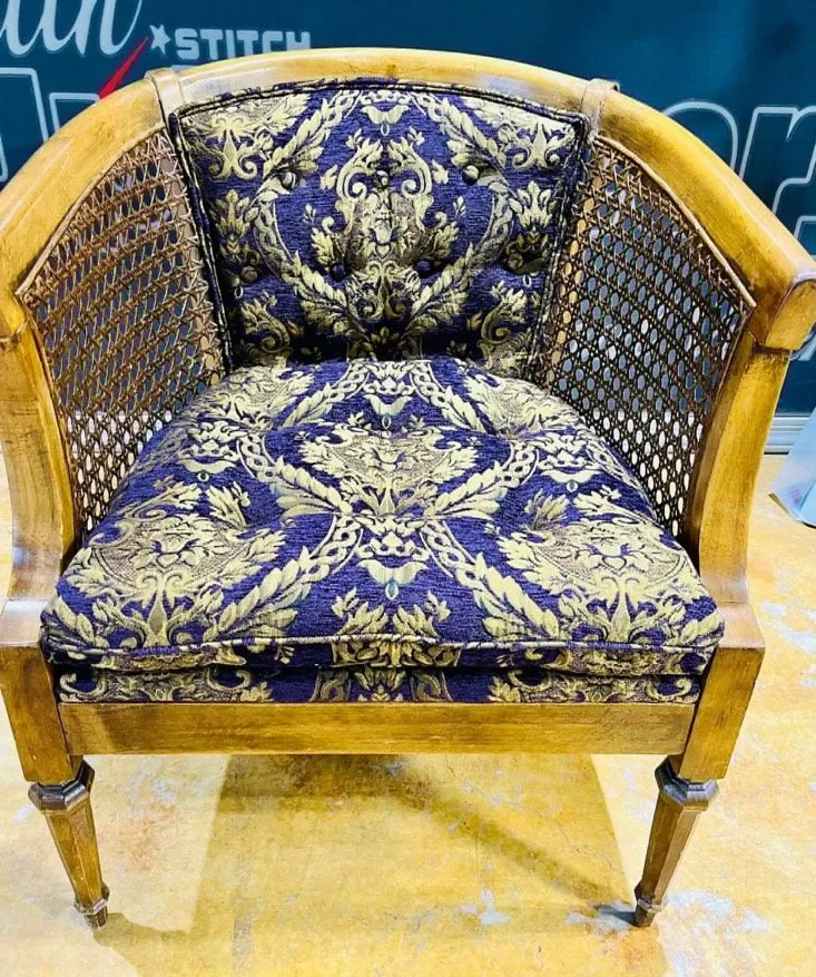 A vintage wooden loveseat with a carved wooden frame and patterned dark blue and gold upholstery, featuring floral and leaf designs.