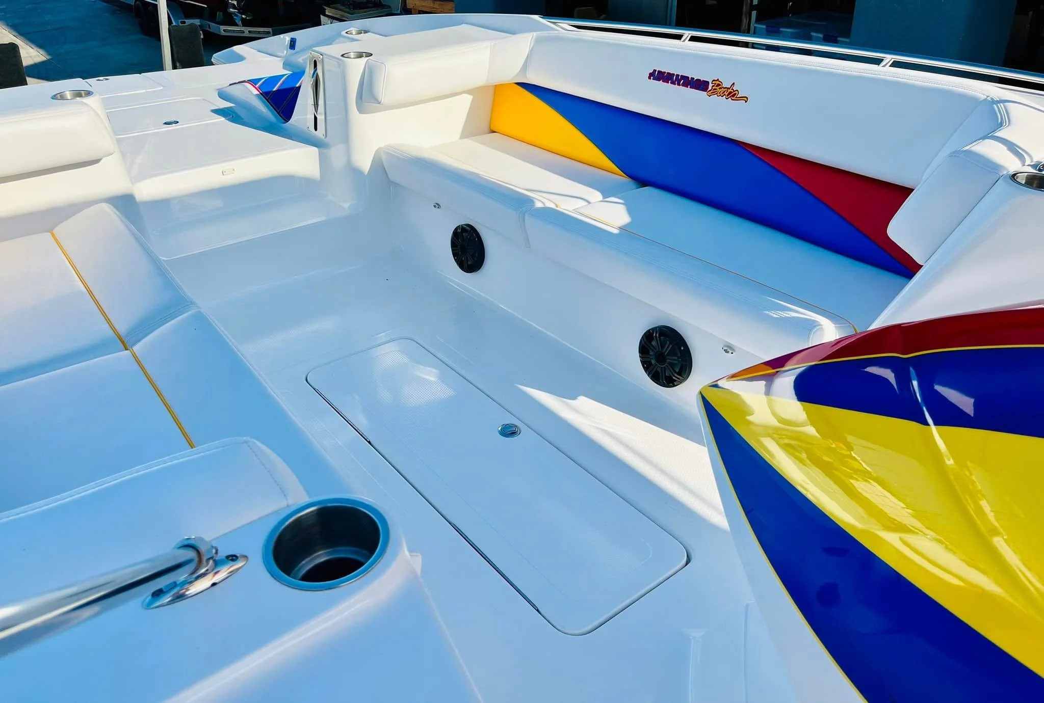 Interior of a speedboat with white seating, a colorful padded backrest with yellow, blue, and red sections, and built-in speakers. A small cup holder and storage compartment are visible. The boat is docked outdoors.