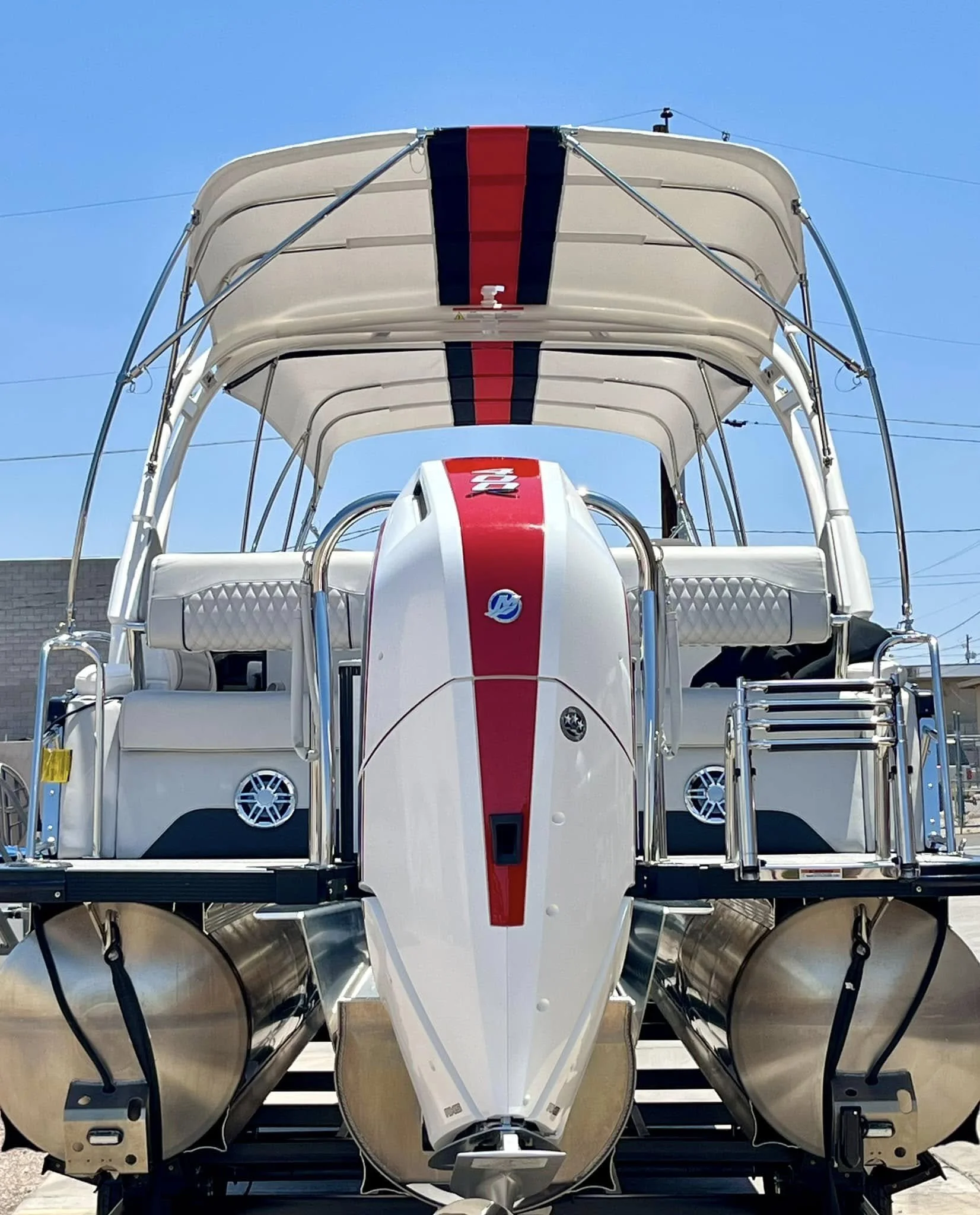 A white pontoon boat with a red and black stripe, metal railings, cushioned seats, a canopy, and dual outboard motors, parked outdoors under a blue sky.