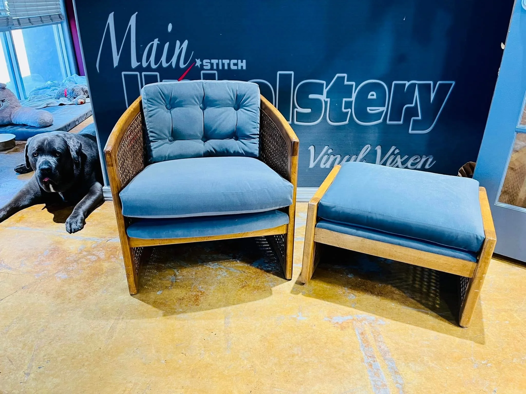 A black dog laying next to a blue cushioned armchair and a matching footstool.