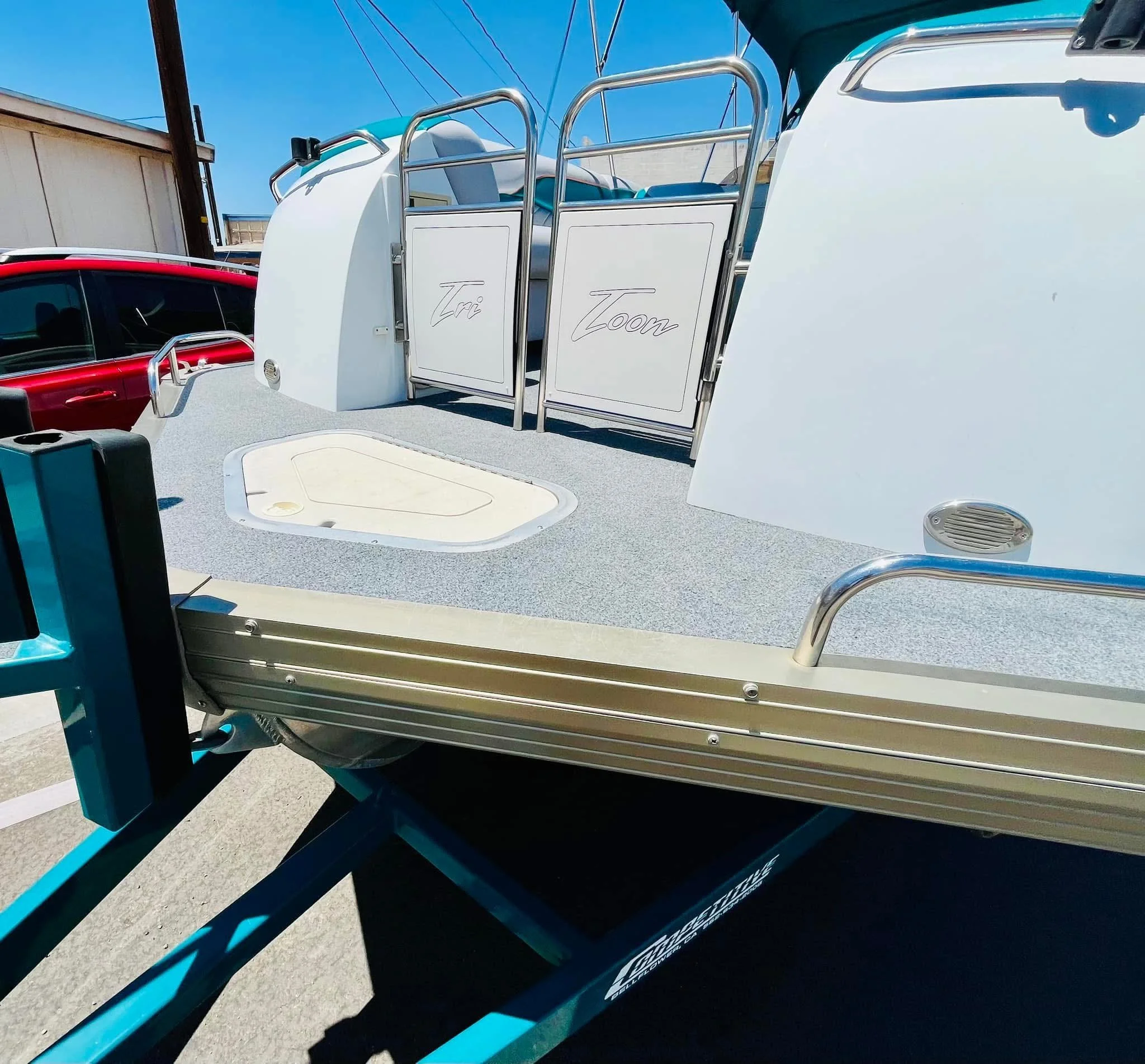 A white and teal boat on a trailer with a red car in the background under a clear blue sky.
