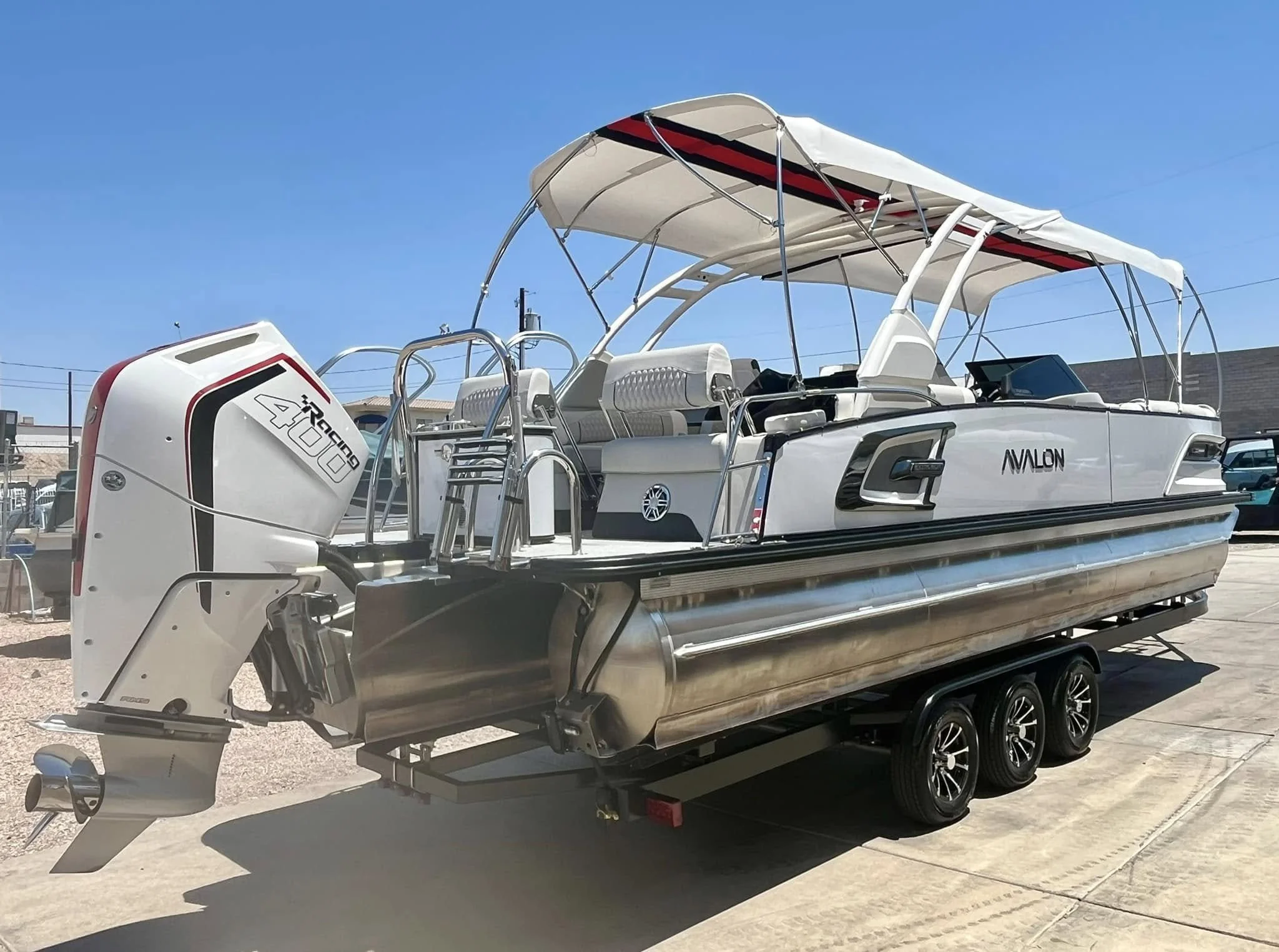 A white Avalon pontoon boat with a black and red canopy, mounted on a black trailer, and attached to a white outboard motor, parked on a concrete surface under a clear blue sky.
