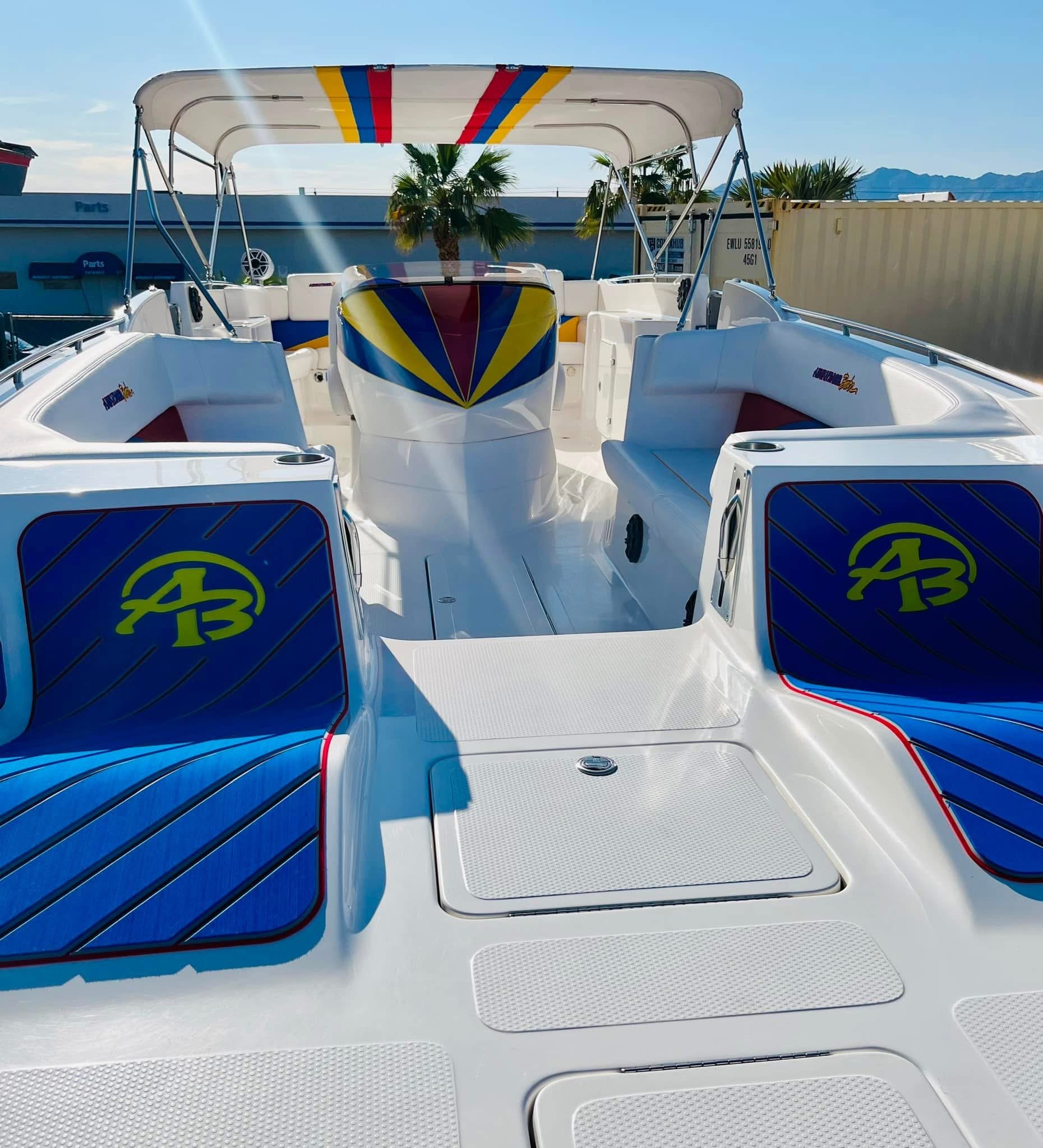 A colorful boat with blue and yellow accents, white seating, and a canopy, parked near palm trees and industrial buildings, with mountains in the background.