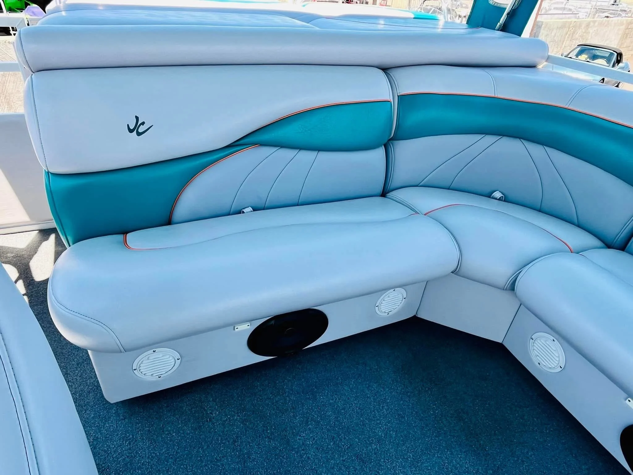 Light blue and teal yacht or boat seating with orange piping, built-in speakers and a black circular speaker, located near a window with visible cars outdoors.