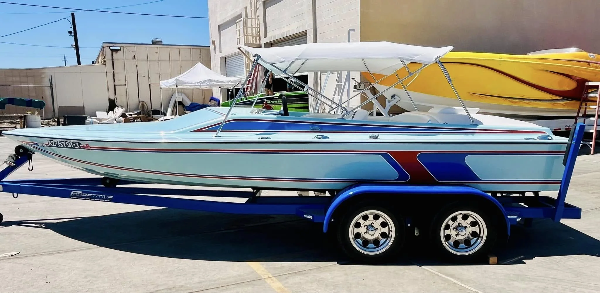 A speedboat with a blue and red striped design on a trailer in an outdoor lot, with other boats and a building in the background.