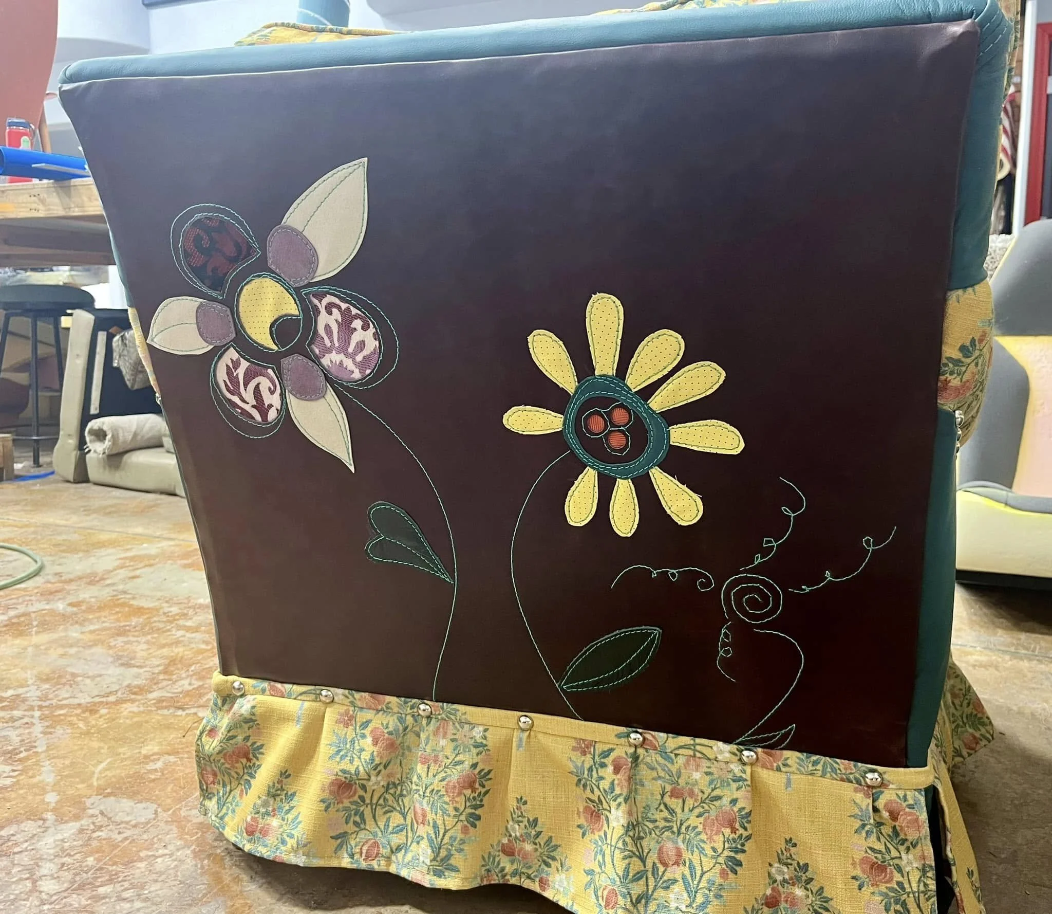 Upholstered furniture with embroidered floral design on the back, featuring a two-tone flower with large petals and black button center, on a yellow fabric skirt with a floral pattern.