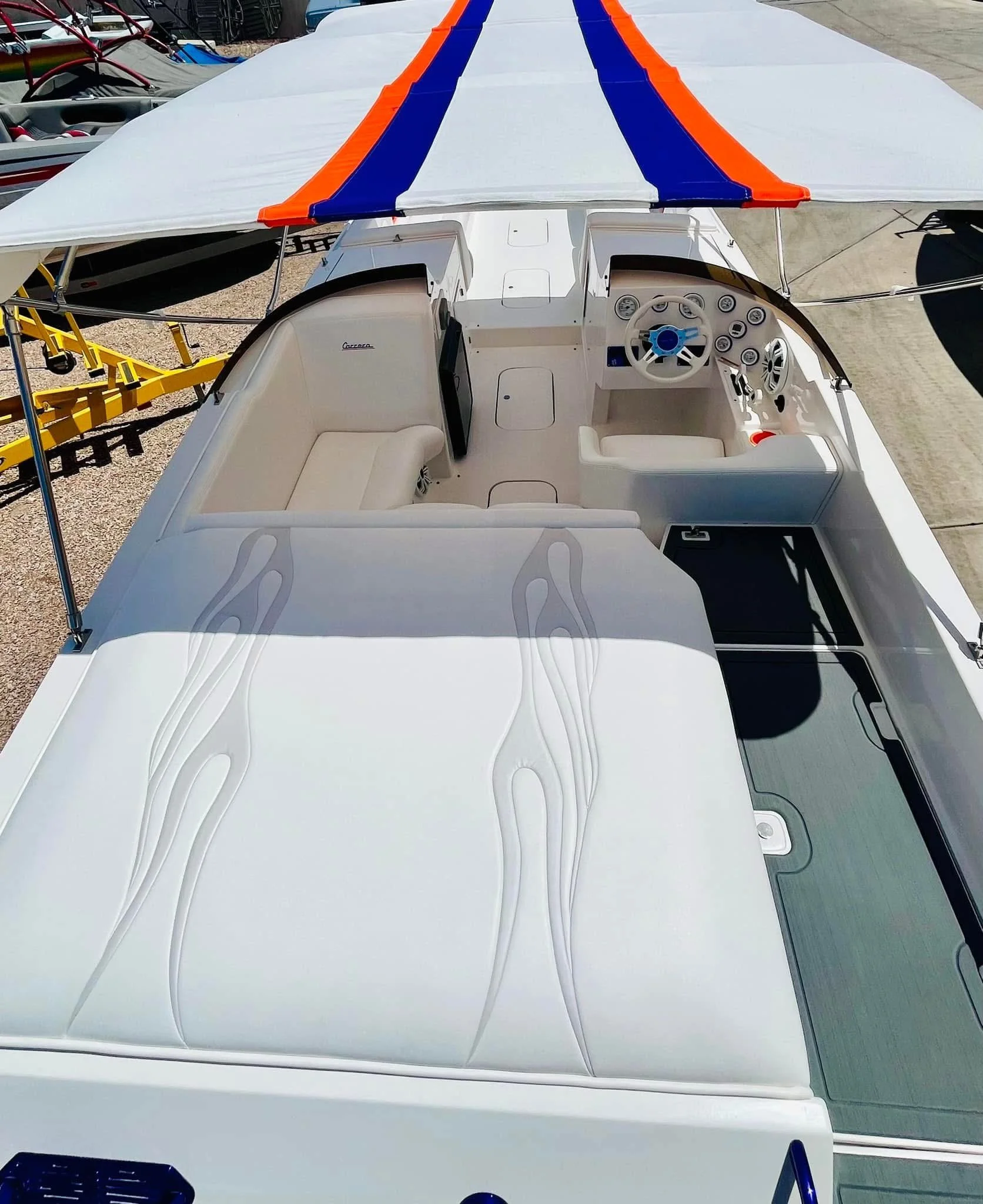 Top view of a white speedboat with a multicolored canopy, featuring seating and a steering wheel, docked near other boats on a concrete surface.