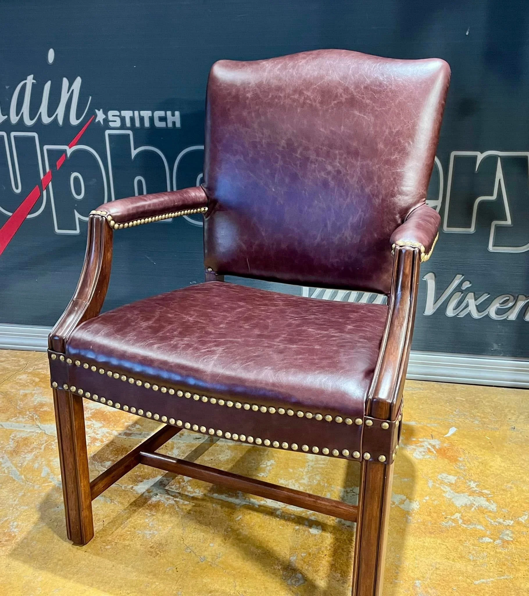 A vintage wooden armchair with red leather upholstery, brass nailhead trim, and a cushioned backrest, positioned on a yellowish floor with a dark wall in the background.