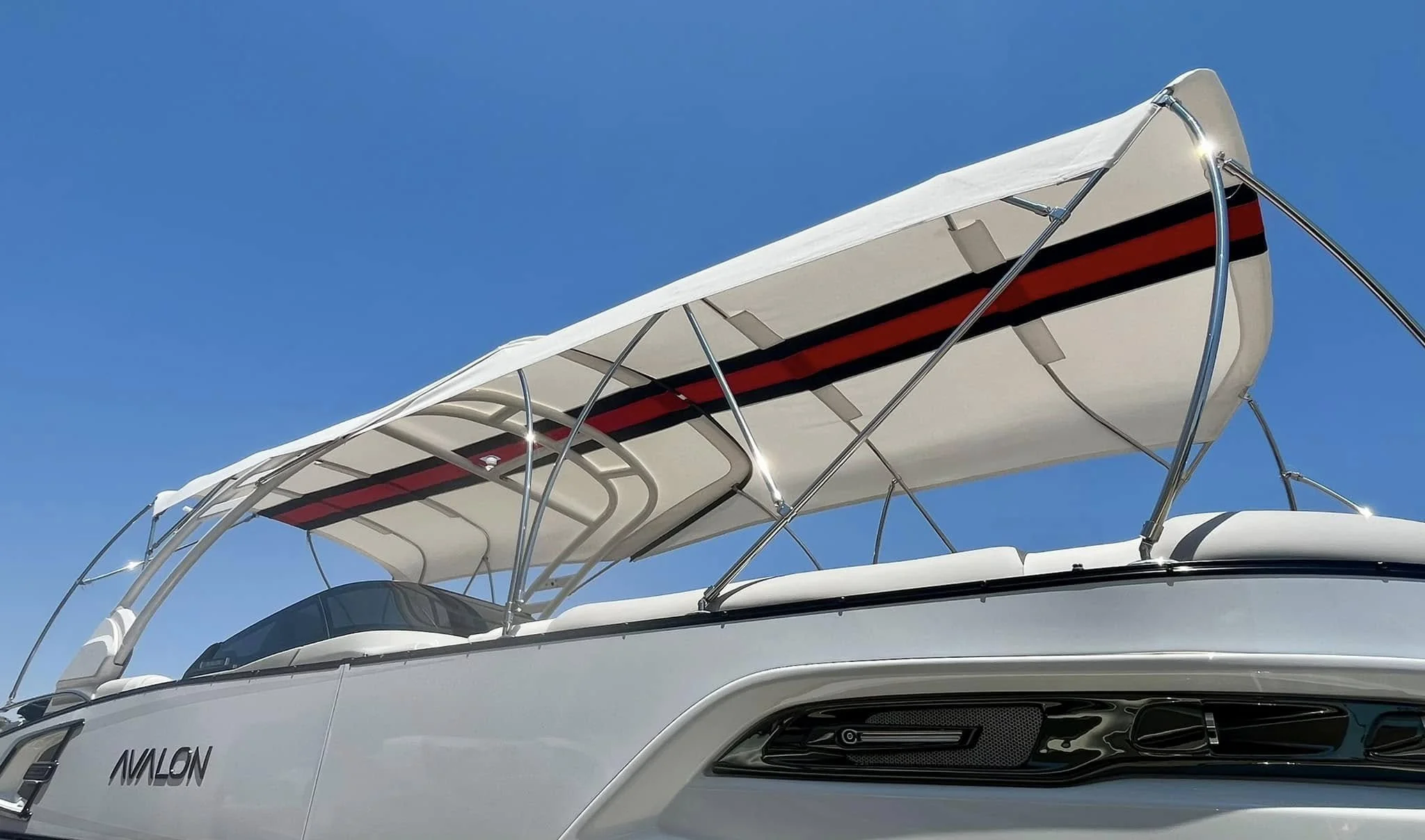 A white Avalon boat with a white canopy and red and black stripe, anchored under a clear blue sky.