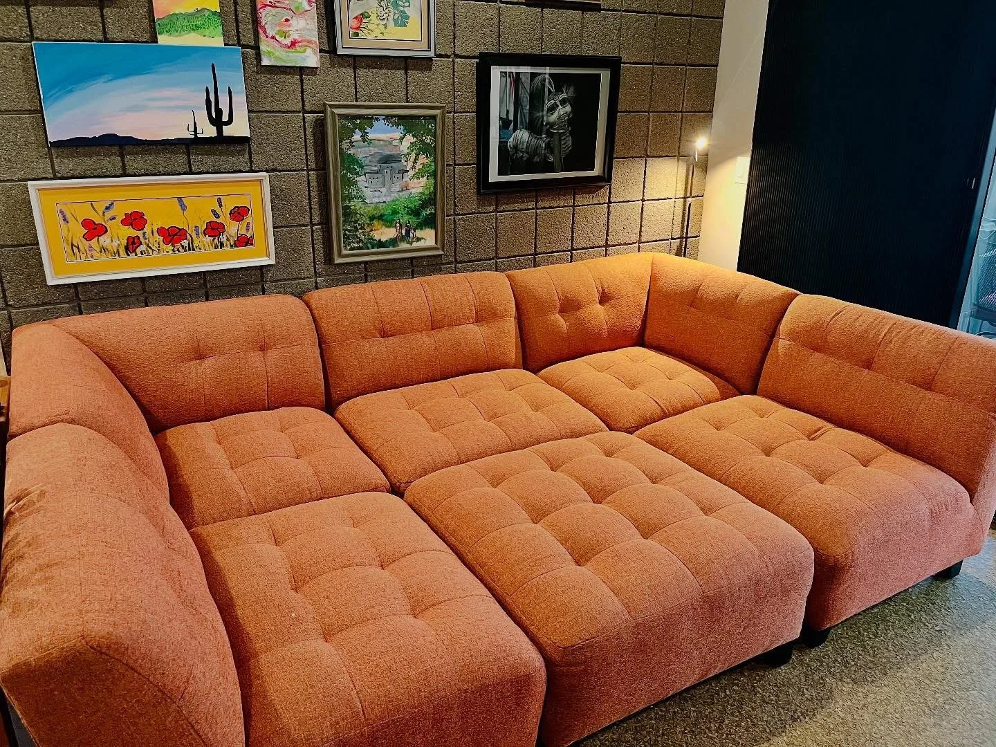 An orange sectional sofa in front of a wall with various framed and unframed colorful artwork, including paintings of cacti, flowers, a landscape, and a skull with sunglasses. There is a lamp and a tall light behind the sofa.