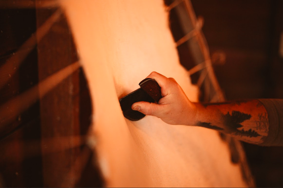 Hand holding single bevel head knife  scraping stretched rawhide on frame.